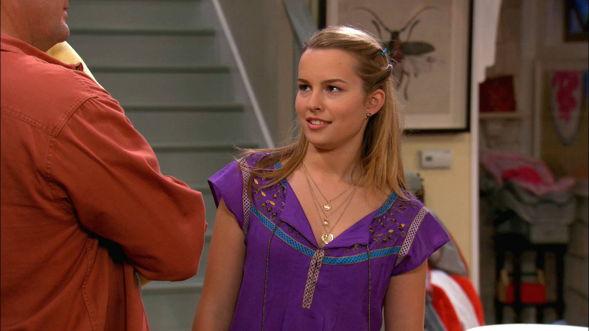 Good Luck Charlie 泰迪·鄧肯 壁紙 — 一位身穿紫色圖案上衣的年輕女性微笑著站在溫暖照明的室內環境中，身後有樓梯和裱框藝術品。