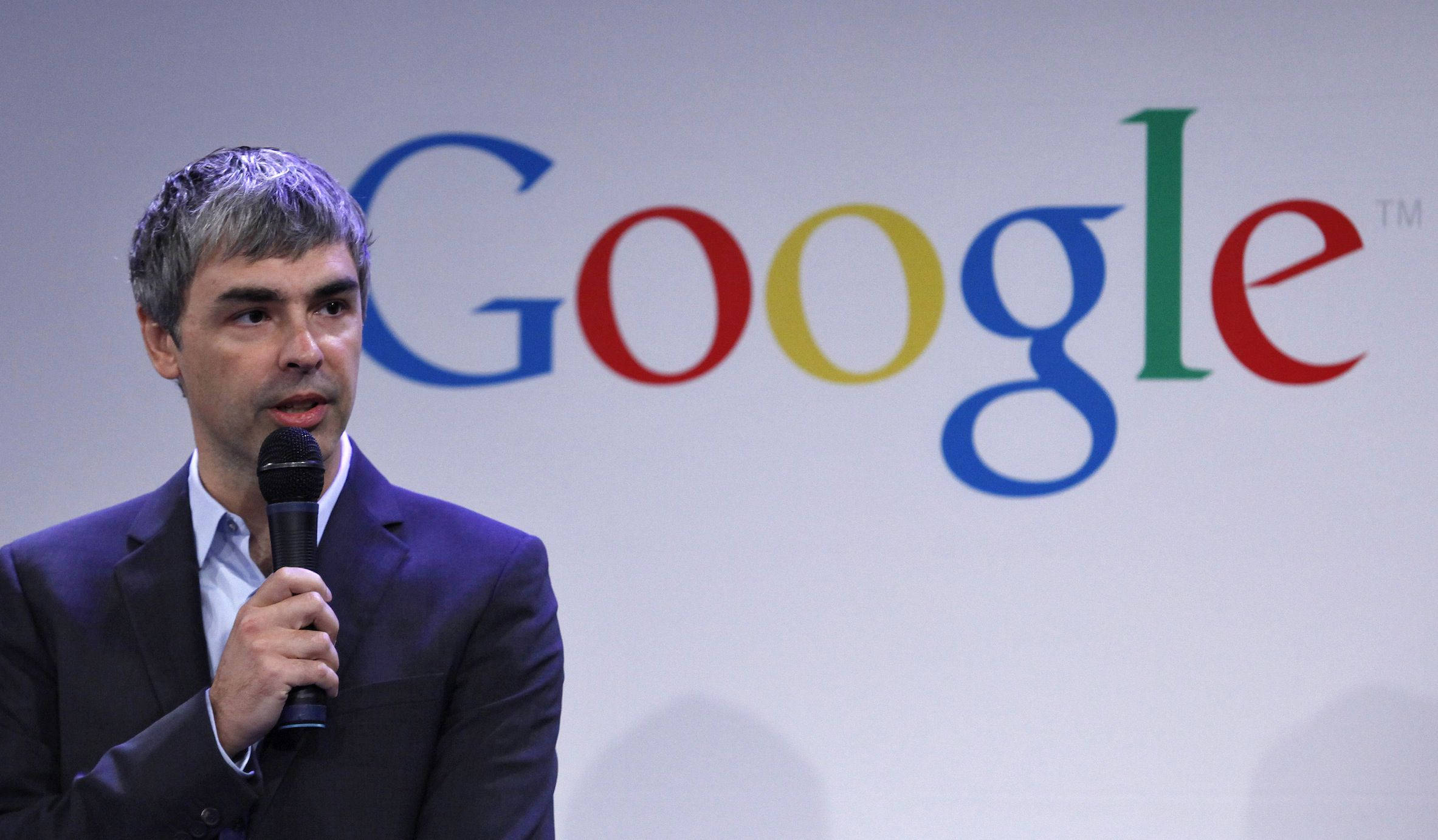 拉里·佩奇Google會議演講攝影 壁紙 — 一名身穿深色西裝的男子站在色彩繽紛的大型Google標誌背景前，手持麥克風。