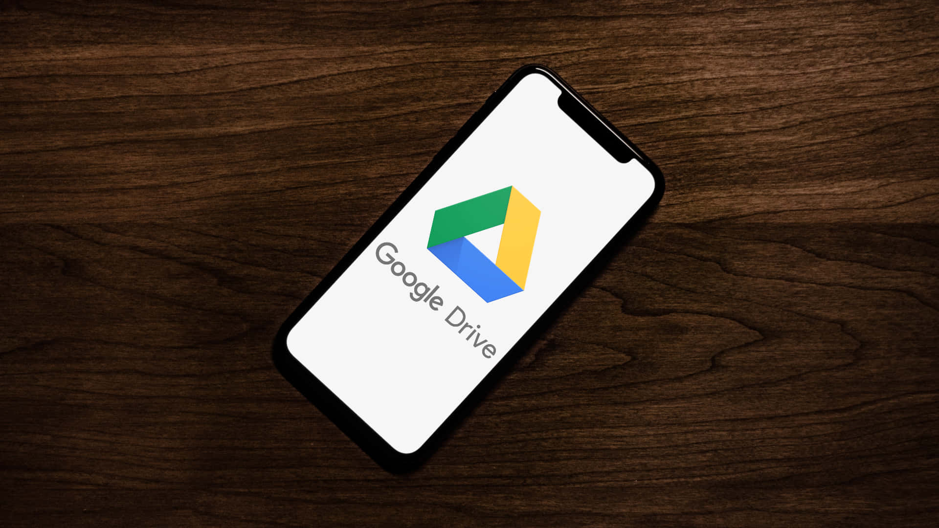 木桌上的 Google Drive 手機介面 壁紙 — 一部顯示Google Drive標誌的智能手機對角放置在深色木質表面上。