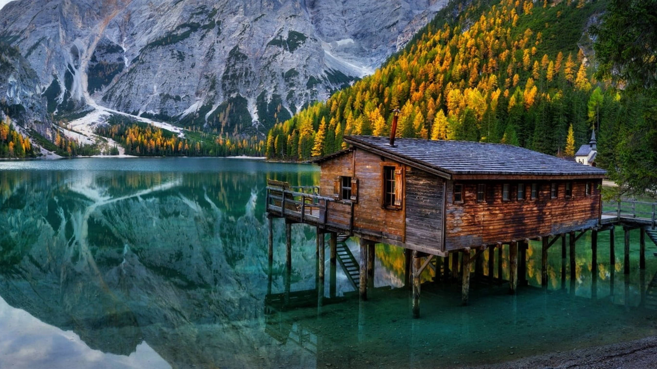 湖中木屋 Google Meet 背景 壁紙 — 一座質樸的木製高腳小屋完美地倒映在清澈的阿爾卑斯湖中，周圍環繞著秋色森林和高聳的岩石山脈。