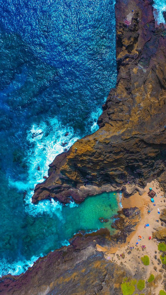 Google Pixel 3 海崖 壁紙 — 俯瞰圖：碧綠海水拍打崎嶇懸崖，環繞一個僻靜沙灘，沙灘上散落著遊客。