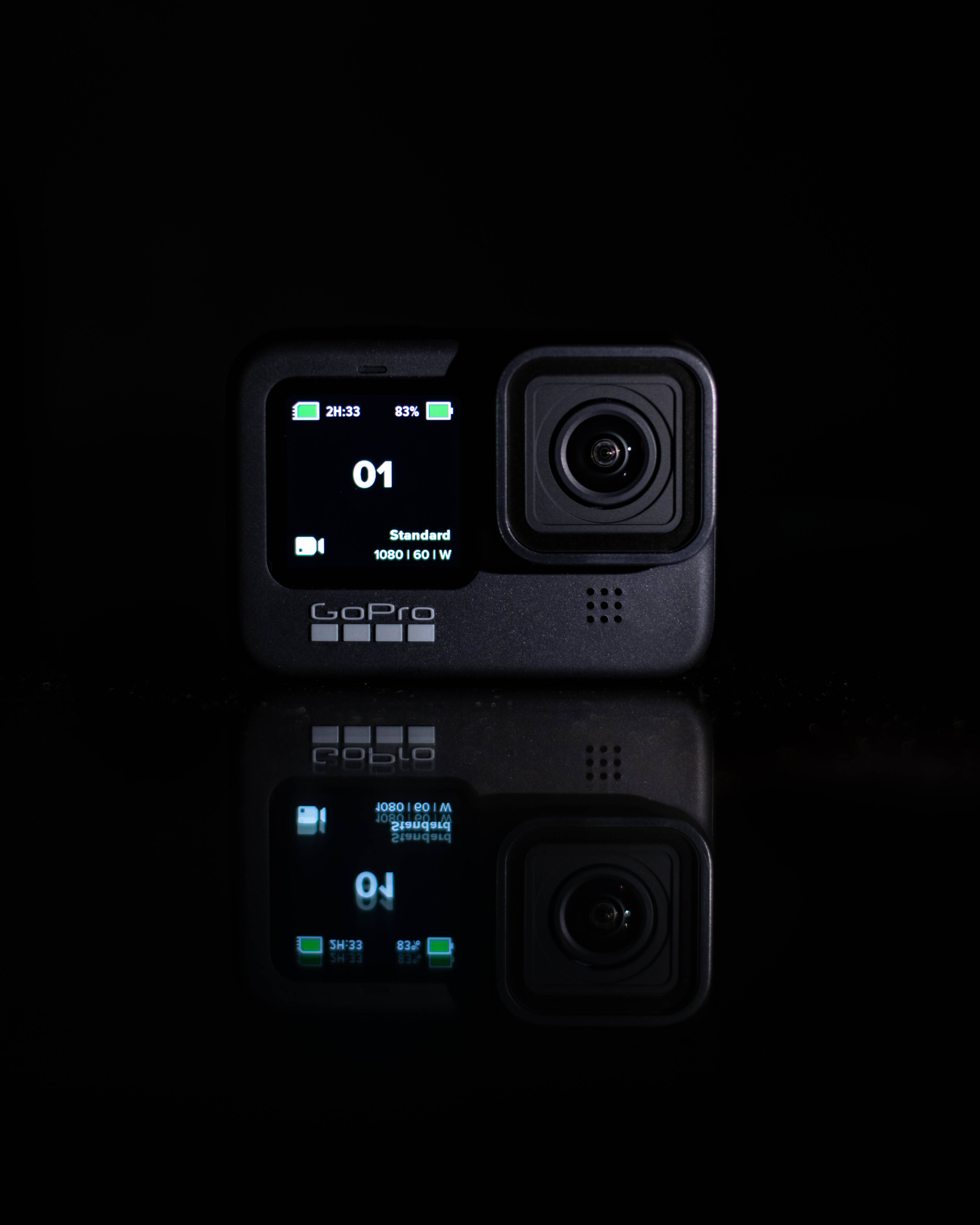 黑色盒型GoPro攝影機 壁紙 — 一部黑色GoPro相機帶有發光綠色螢幕細節，放置在反射表面上，背景為深色。