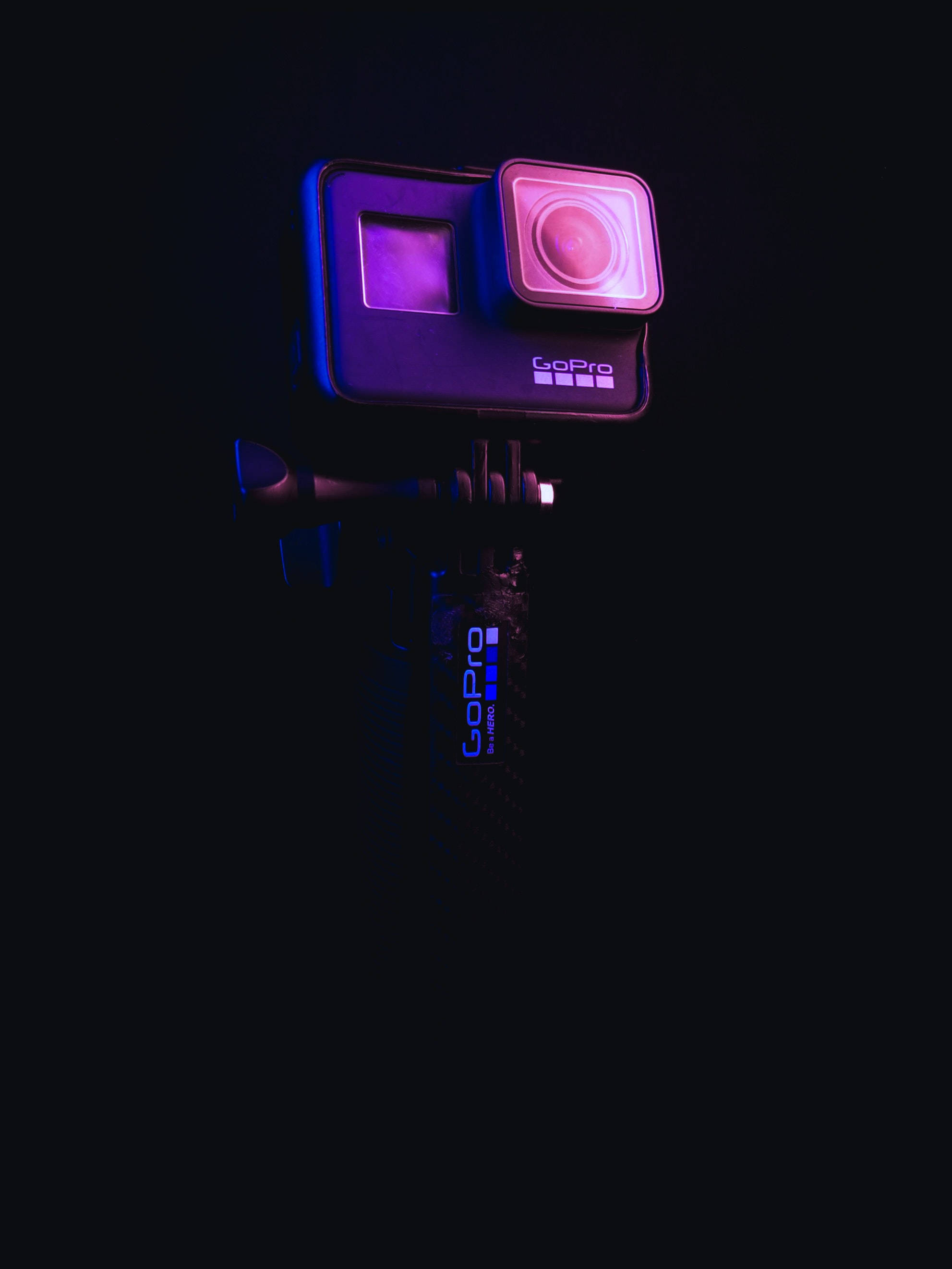 GoPro 在霓虹紫光下 壁紙 — 一部安裝在黑色三腳架臂上的GoPro相機，在漆黑背景下散發出鮮豔的紫色和藍色霓虹燈光。