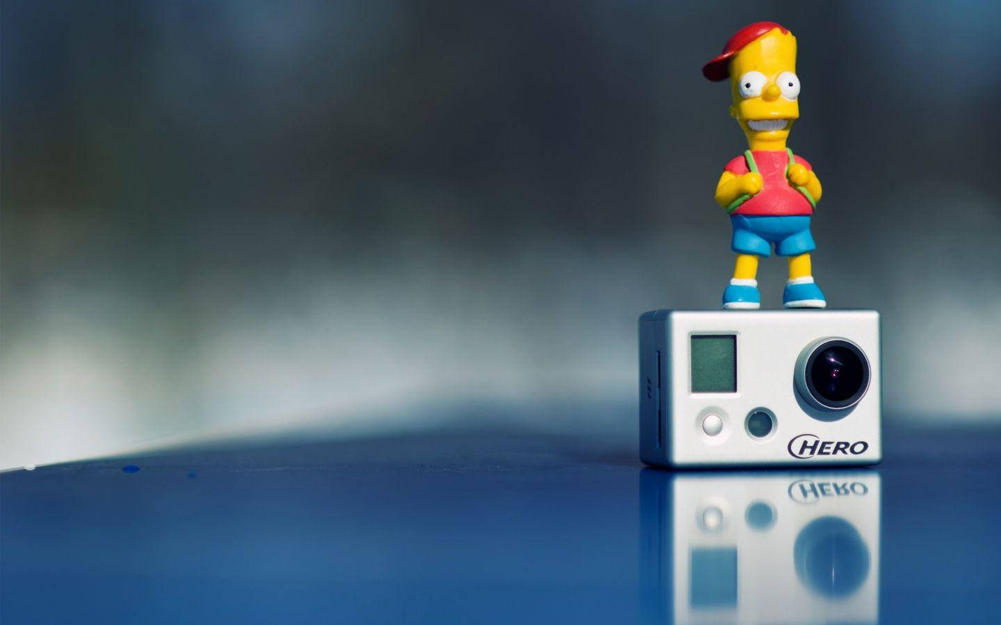 GoPro 巴特·辛普森小人偶 壁紙 — 一個巴特·辛普森公仔企喺白色GoPro HERO相機上面，喺一塊反光藍色表面上，背景模糊。