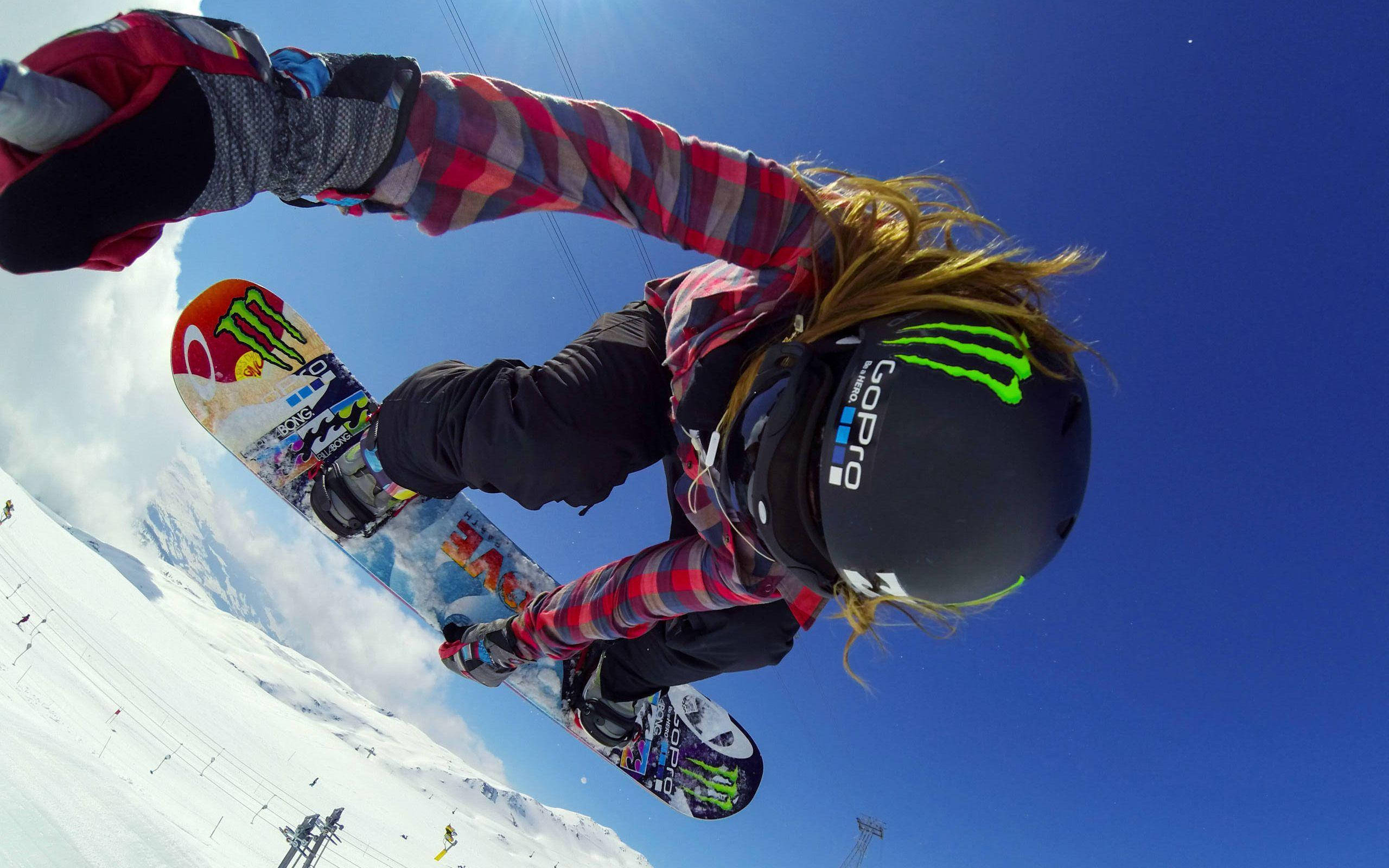 GoPro 拍攝的滑雪板動作瞬間 壁紙 — 一名滑雪板運動員在明亮的藍天下騰空，下方是白雪覆蓋的山脈。