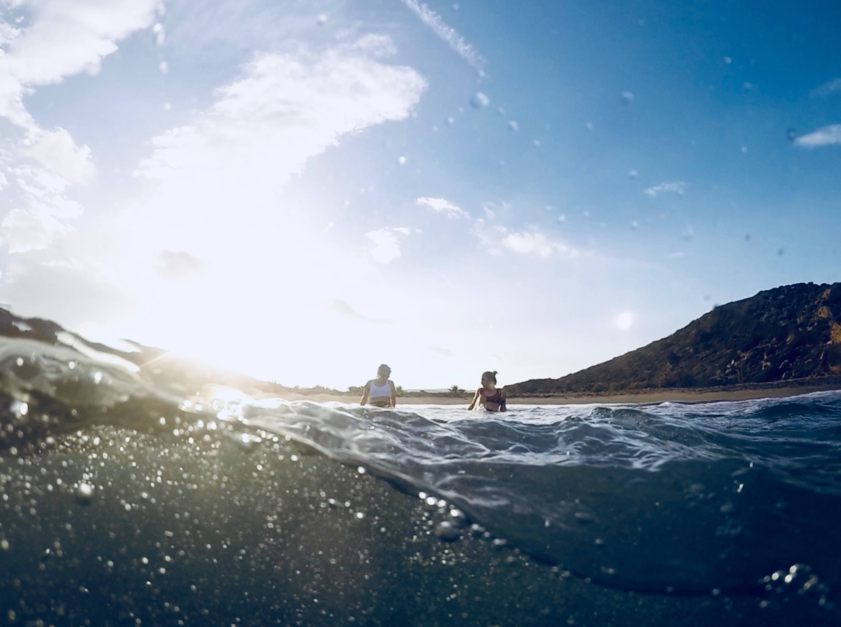 GoPro 拍攝的衝浪照片 壁紙 — 兩個人在燦爛陽光下涉水走過海浪，背景有山丘，從水下視角觀看。
