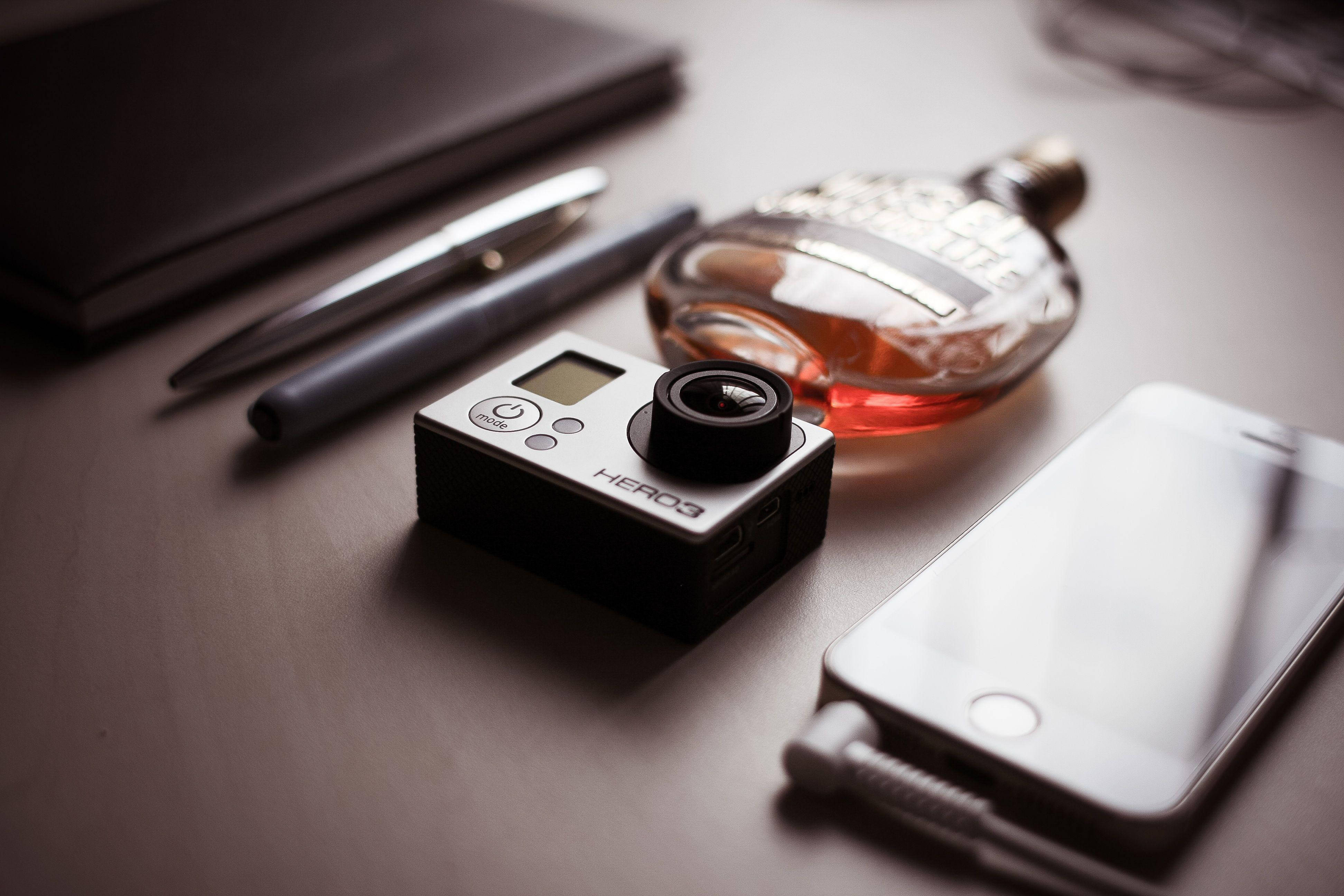 GoPro 與香水瓶 壁紙 — 一部時尚的GoPro相機放在智能手機威士忌酒瓶和筆旁邊，置於簡約木桌上。