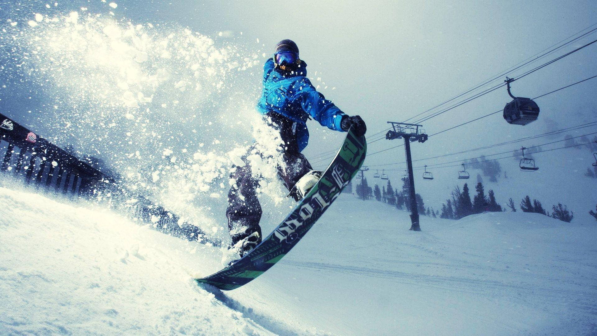 GoPro 雪上極限雪板者 壁紙 — 一名身穿藍色外套的滑雪板運動員在雪坡上轉彎，揚起一陣粉雪，背景有滑雪纜車。