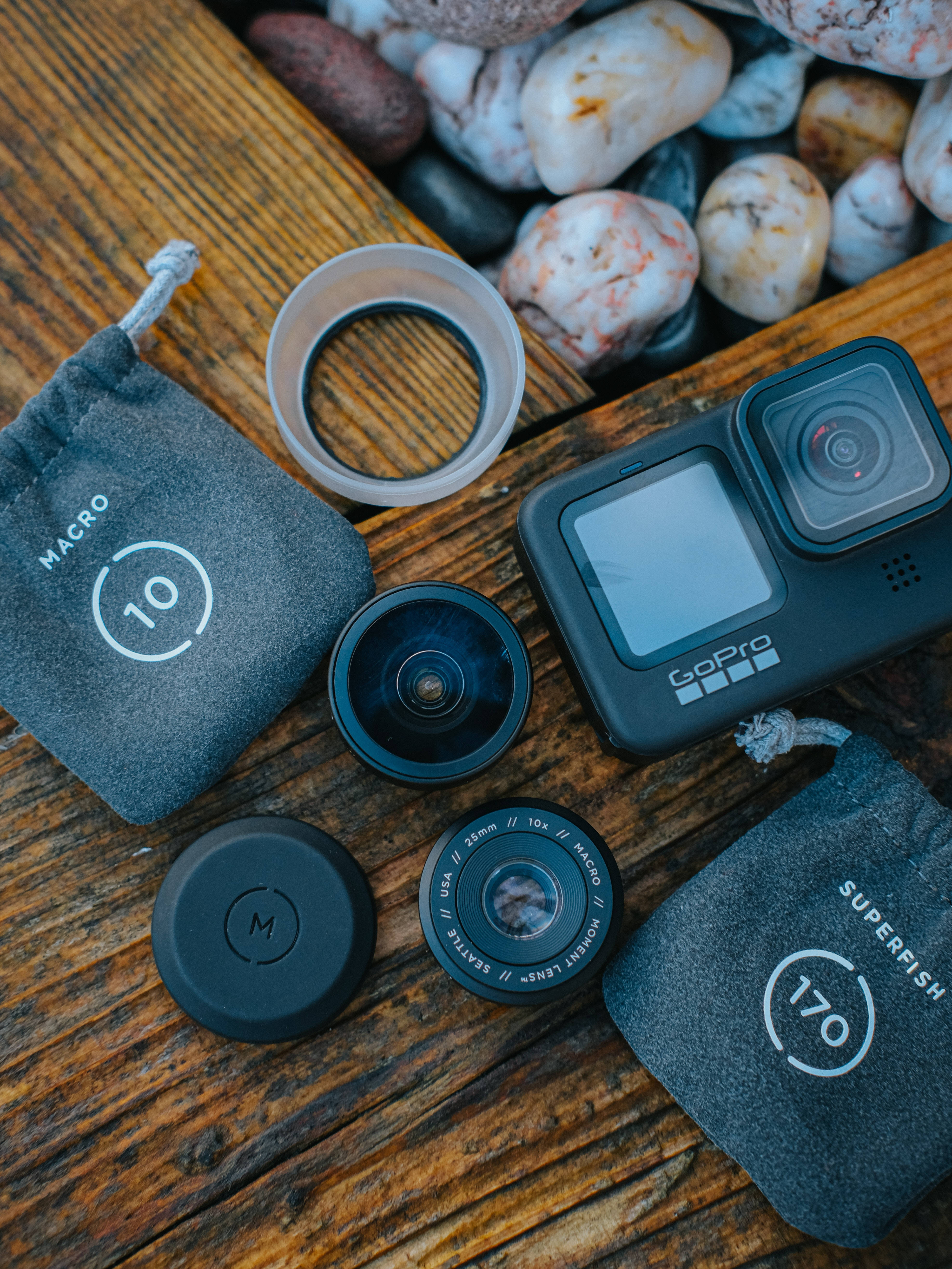GoPro不同鏡頭配件 壁紙 — GoPro相機及其微距超級魚眼鏡頭排列在質樸的木質表面上，旁邊是光滑的河石。