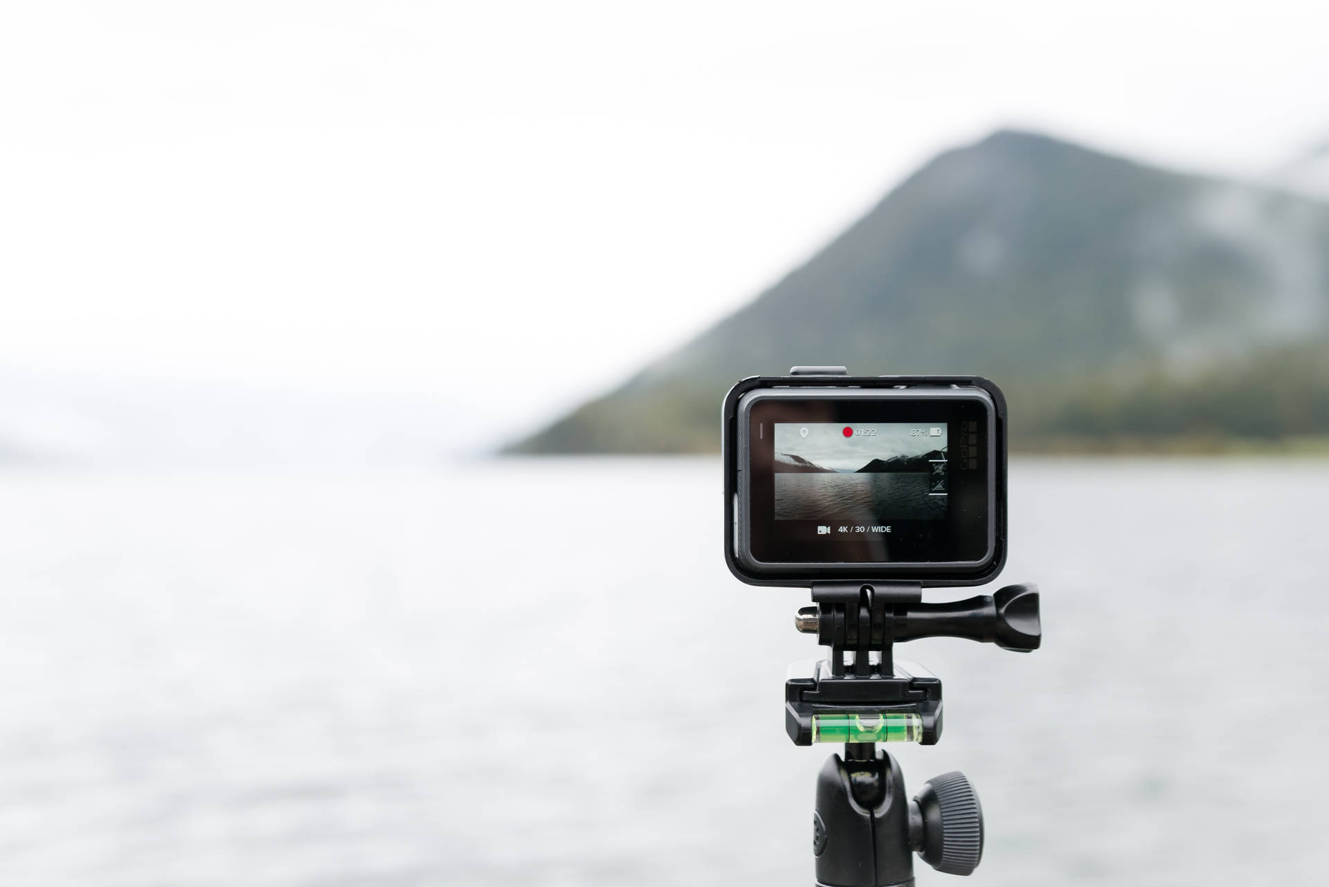 GoPro拍攝島嶼 壁紙 — 黑色運動相機安裝在三腳架上，錄製背景有模糊山脈的霧氣湖泊。