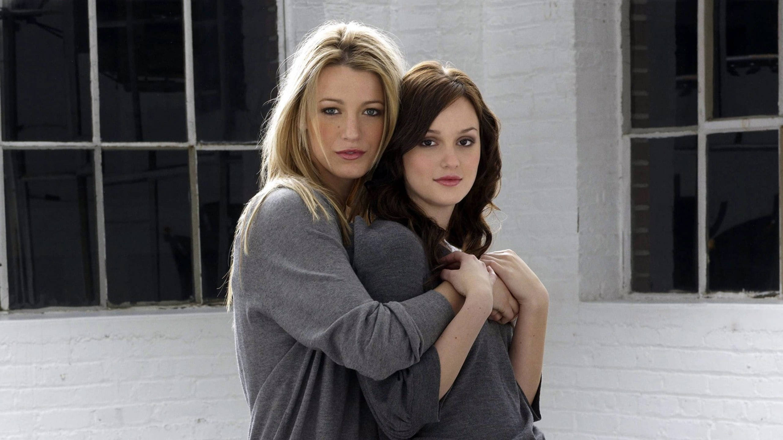 《Gossip Girl》塞雷娜與布萊爾灰色衛衣 壁紙 — 兩位身穿灰色毛衣的女性在白色磚牆和大型深色窗戶前溫暖擁抱。