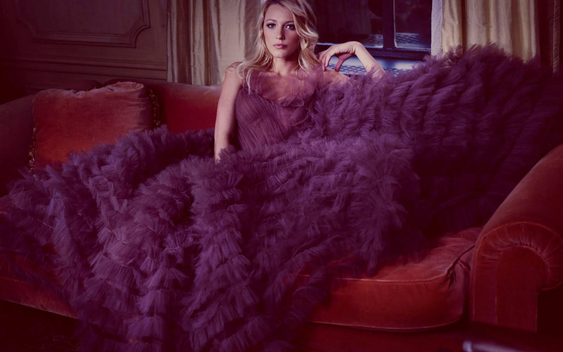 《Gossip Girl》賽琳娜穿絨毛禮服 壁紙 — 一位身穿紫色羽毛長裙的女性，端莊地坐在光線昏暗的優雅房間裡的柔軟紅色天鵝絨沙發上。