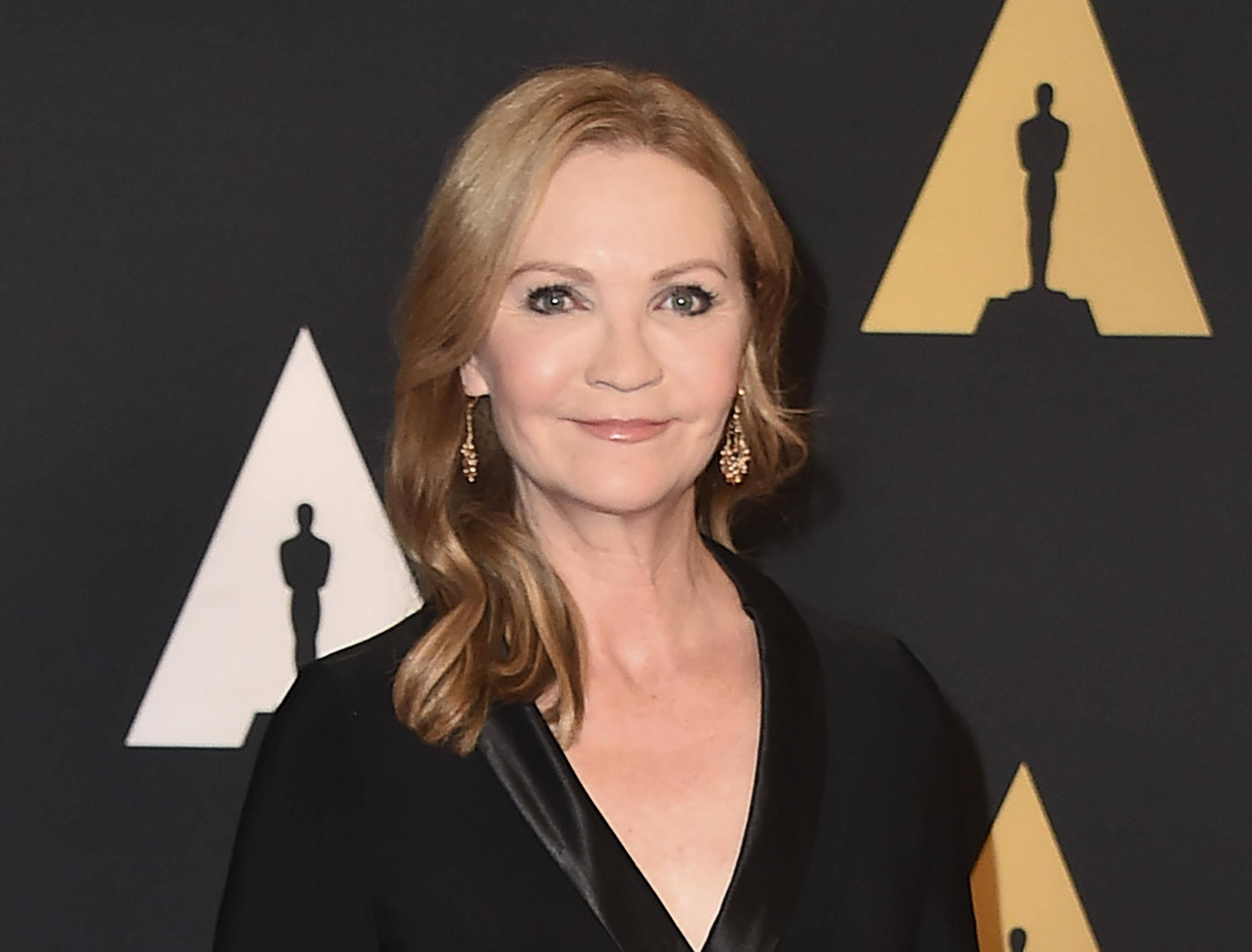瓊·艾倫在 Governors Awards 擔任有聲書朗讀 壁紙 — 一位金髮碧眼、戴著優雅耳環的女性在黑色背景前對著鏡頭微笑，背景上有金色和白色的奧斯卡小金人標誌。