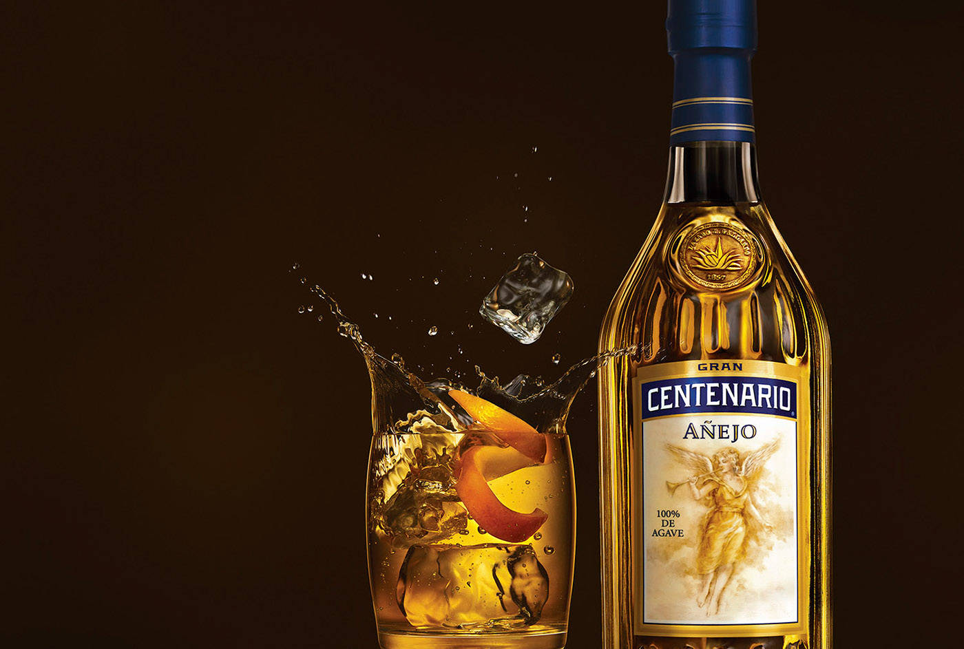 一瓶Centenario Añejo龍舌蘭酒，旁邊有冰塊和柑橘飛濺的玻璃杯。