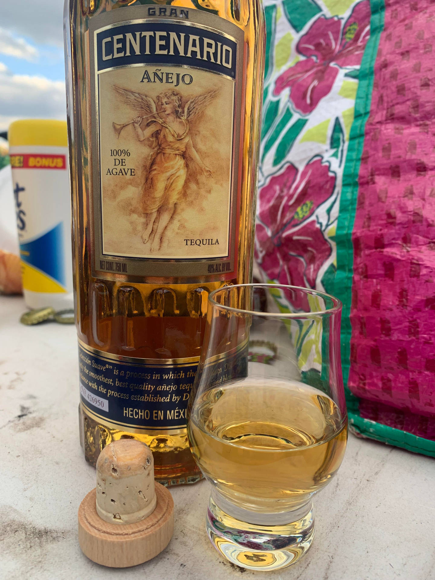一瓶龍舌蘭酒，旁邊有杯子和軟木塞，放在桌子上，旁邊是花卉布料，在晴朗的天空下。