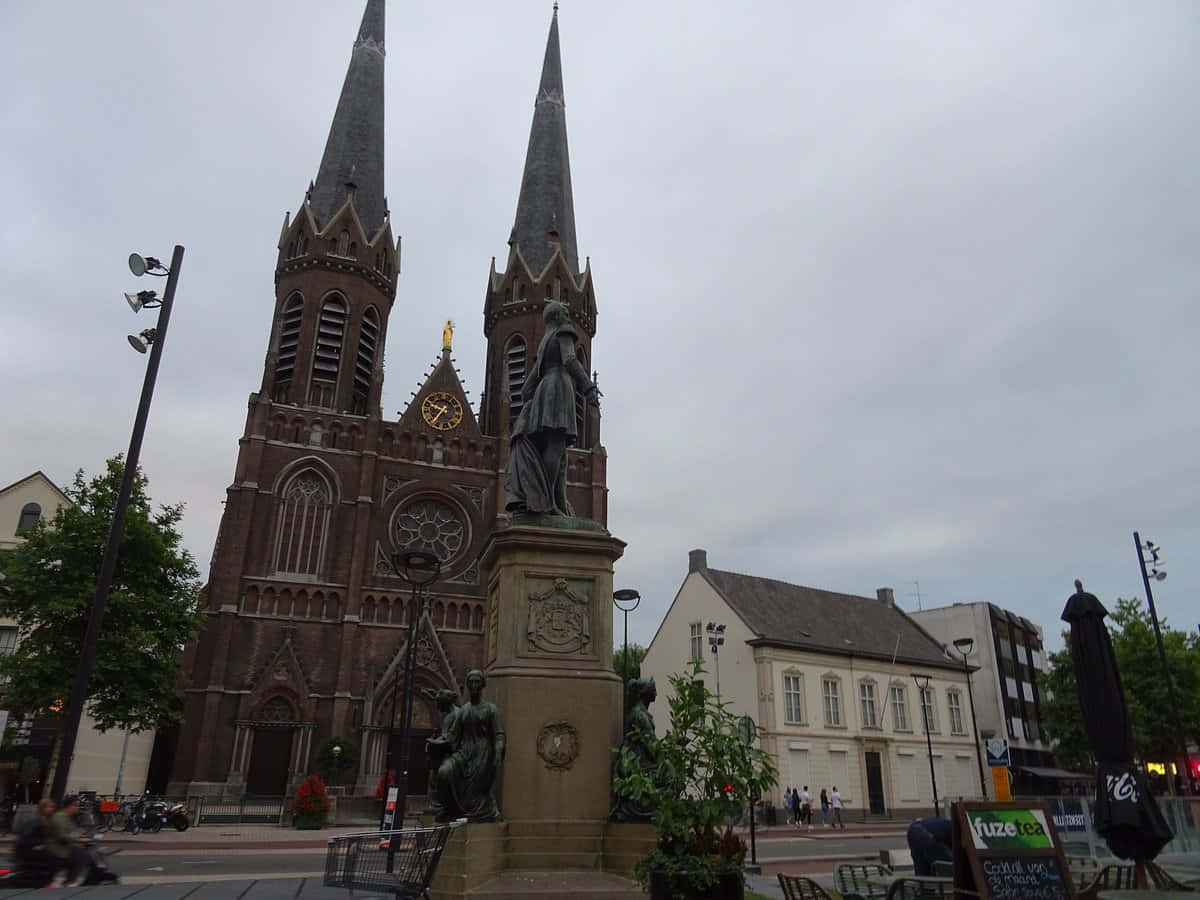Tilburg 桌布 壁紙 — 一座擁有雙尖塔的宏偉哥德式教堂聳立在城市廣場上，廣場中央有雕像和咖啡座，天空多雲。