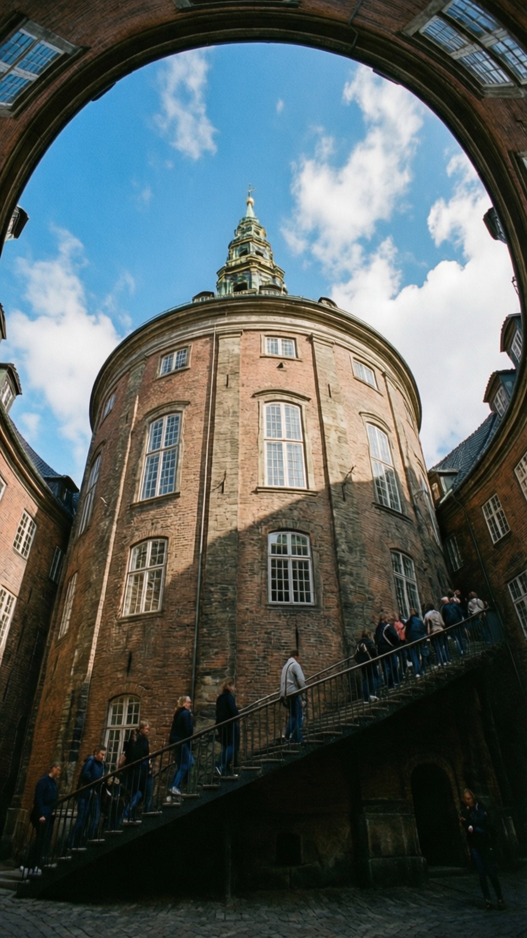 Denmark 桌布 壁紙 — 一座宏偉的歷史磚塔，透過石拱門可見，遊客在明亮的藍天下登上樓梯。
