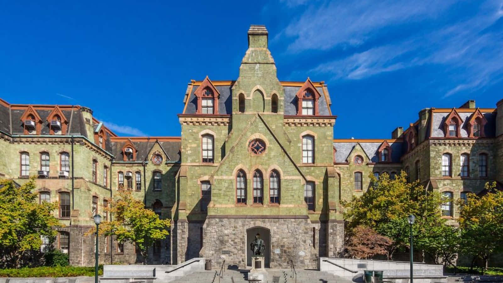  壁紙 — 宏偉的歷史石造大學建築，帶有紅色屋頂點綴和拱形窗戶，背景是清澈的藍天。