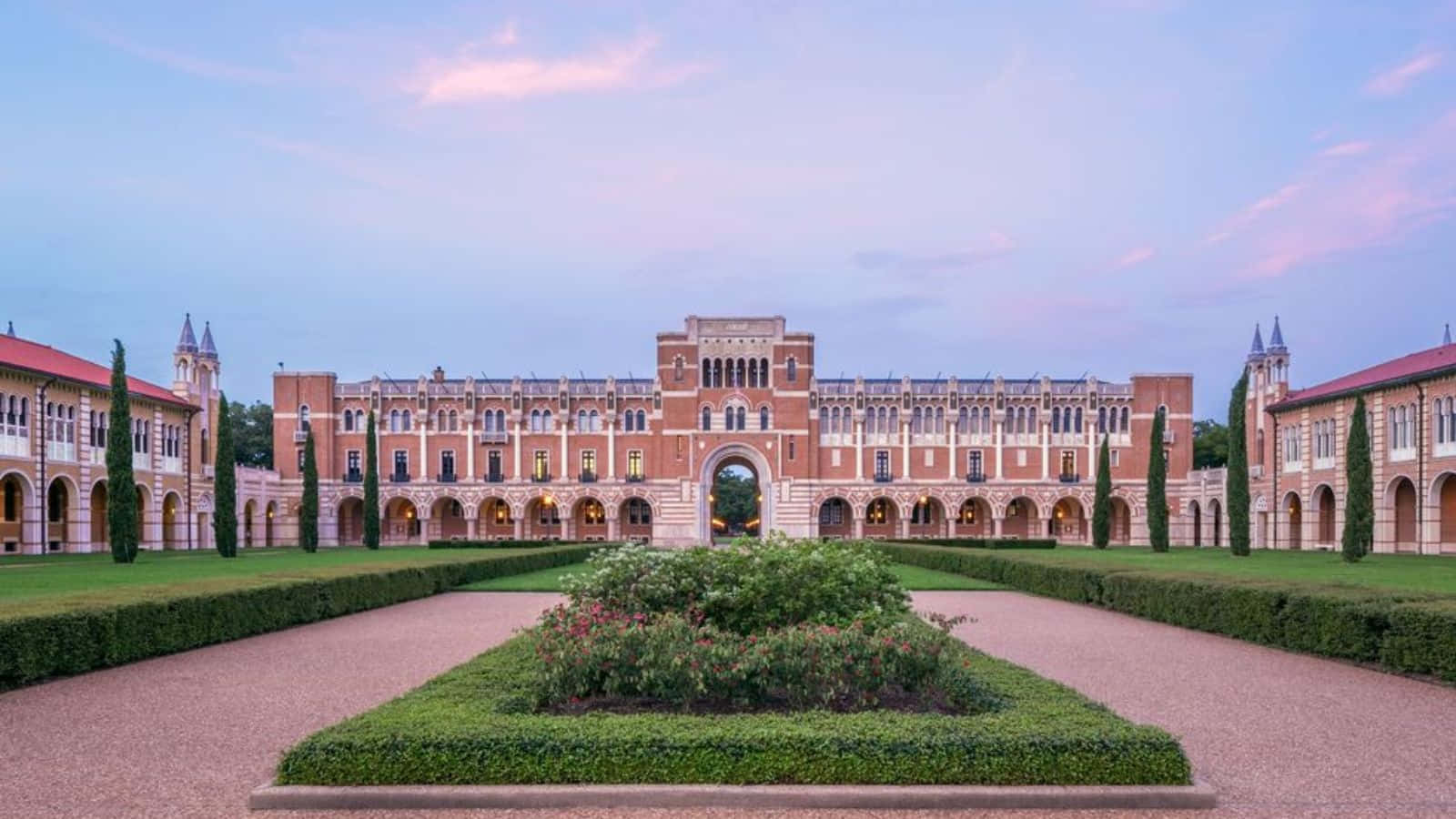 壁紙 — 宏偉的歷史大學建築，紅磚外牆和拱形步道，在暮色天空下由修剪整齊的樹籬框住。