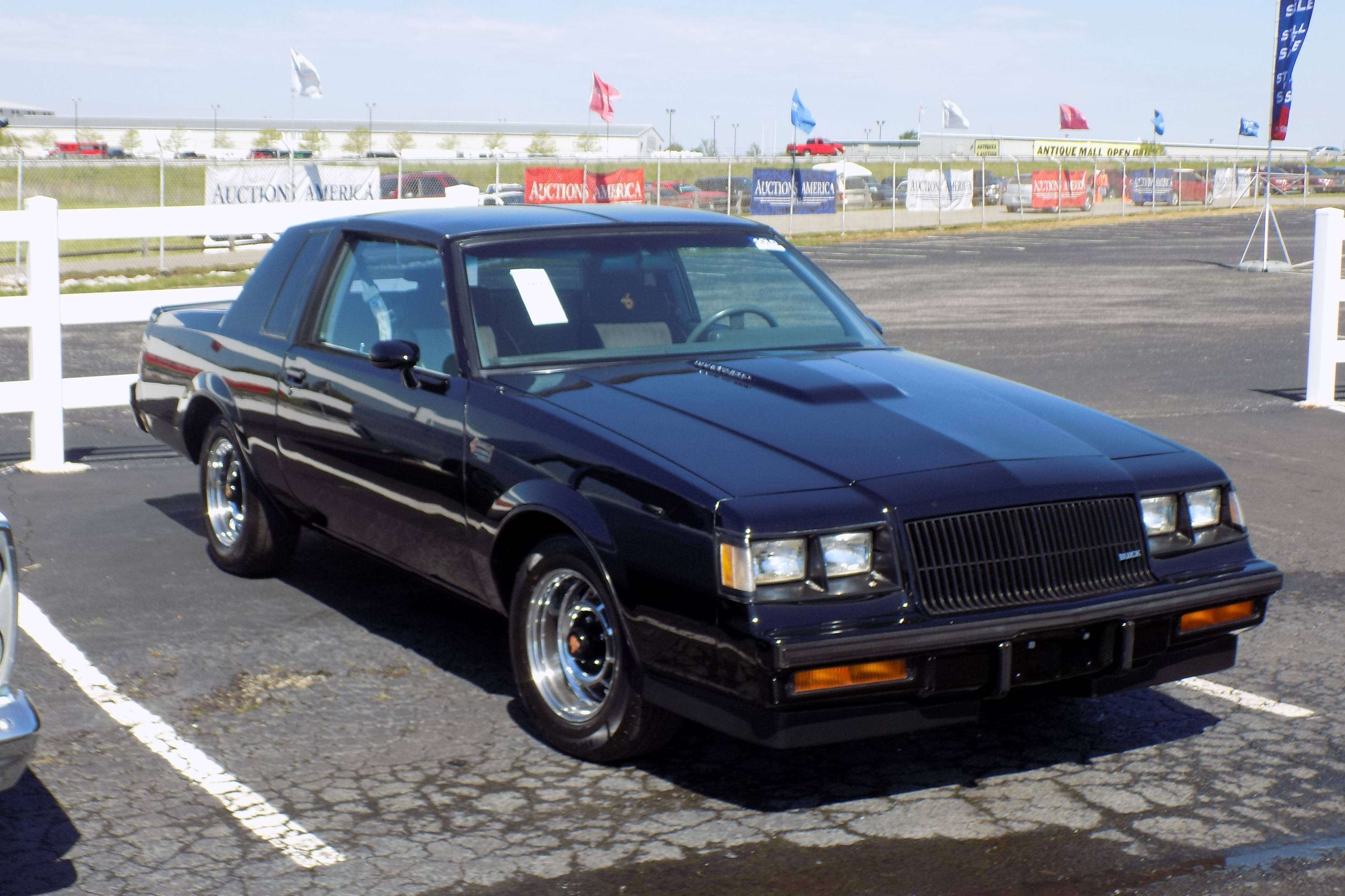 維修良好的別克Grand National 壁紙 — 一輛閃亮的黑色1980年代別克Grand National肌肉車停放在陽光明媚的戶外拍賣場，背景有白色圍欄和旗幟。