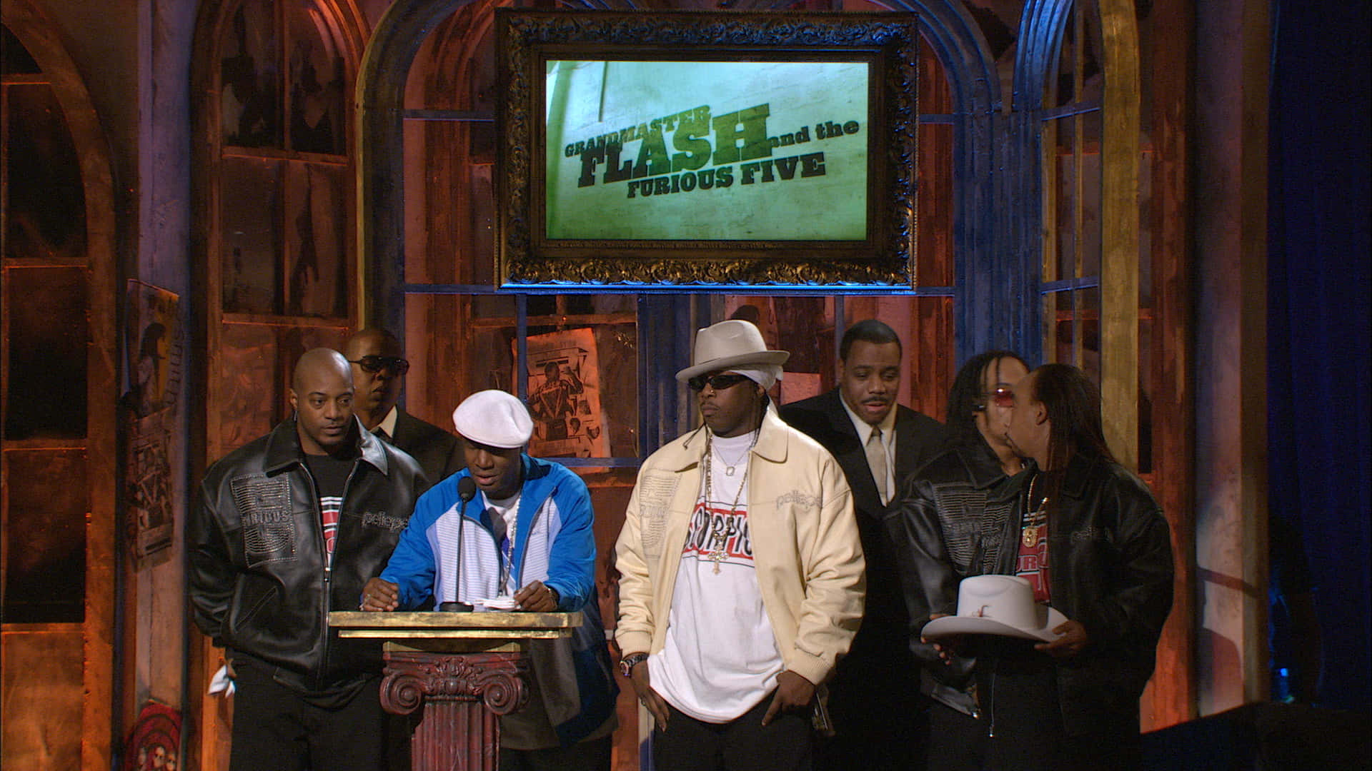 Grandmaster Flash And The Furious Five 三月活動 壁紙 — 五名身穿時尚街頭服飾的男子站在舞台上，身後有一個帶框的屏幕顯示著「Grandmaster Flash and the Furious Five」。