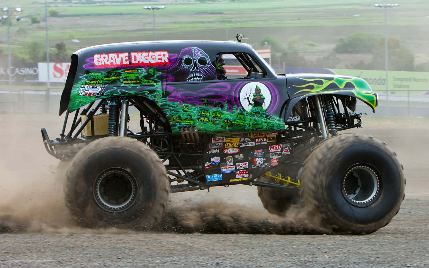 Grave Digger 怪物卡車沙塵飛揚 壁紙 — Grave Digger 怪獸卡車在泥地賽道上咆哮，噴出綠色、紫色和黑色的火焰，揚起塵土。