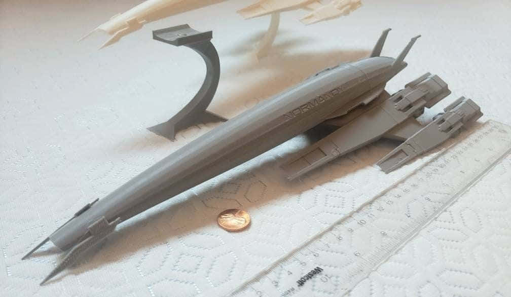 Normandy Sr-2 桌布 壁紙 — 一個灰色3D打印的太空船模型，標有「NORMA」，放置在帶有紋理的白色表面上，旁邊有尺子和硬幣作為比例參考。