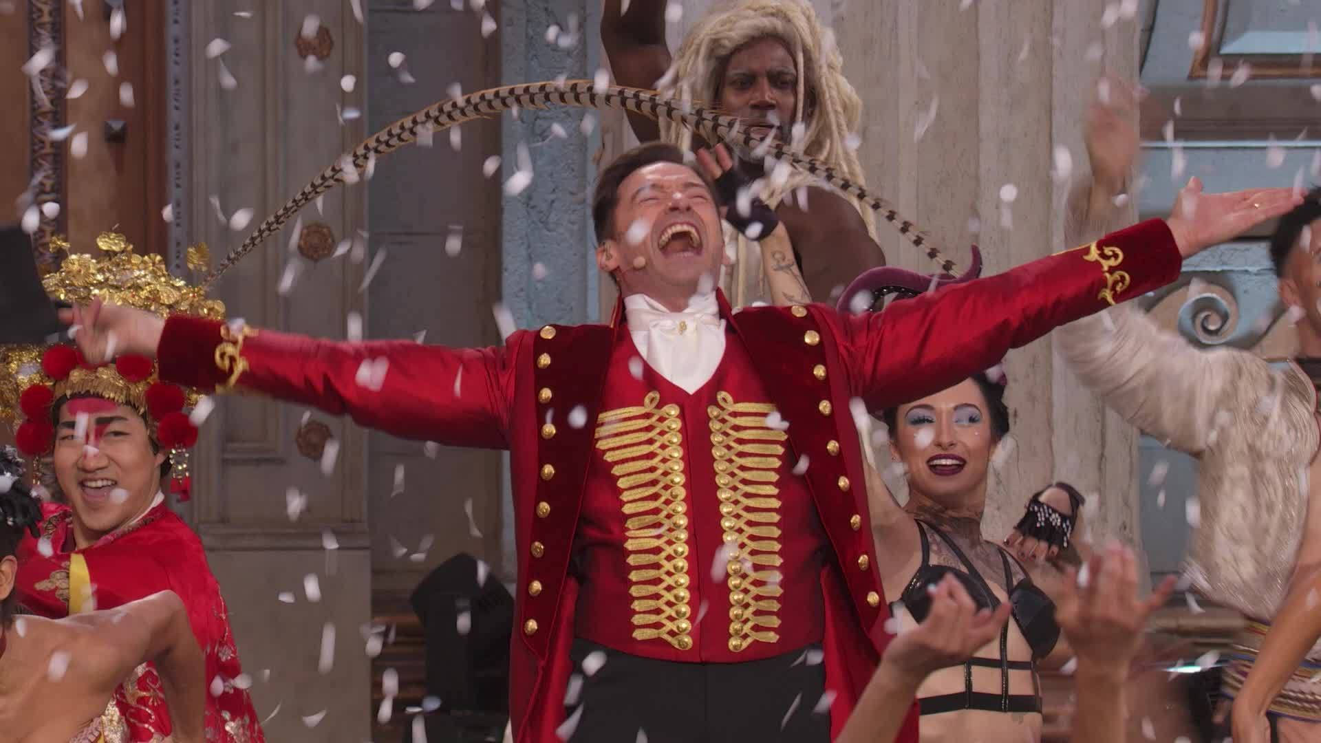 《 Greatest Showman 》精彩演出 壁紙 — 一個快樂的男人穿著紅色和金色的馬戲團服裝，張開雙臂大笑，五彩紙屑在他周圍飄落。