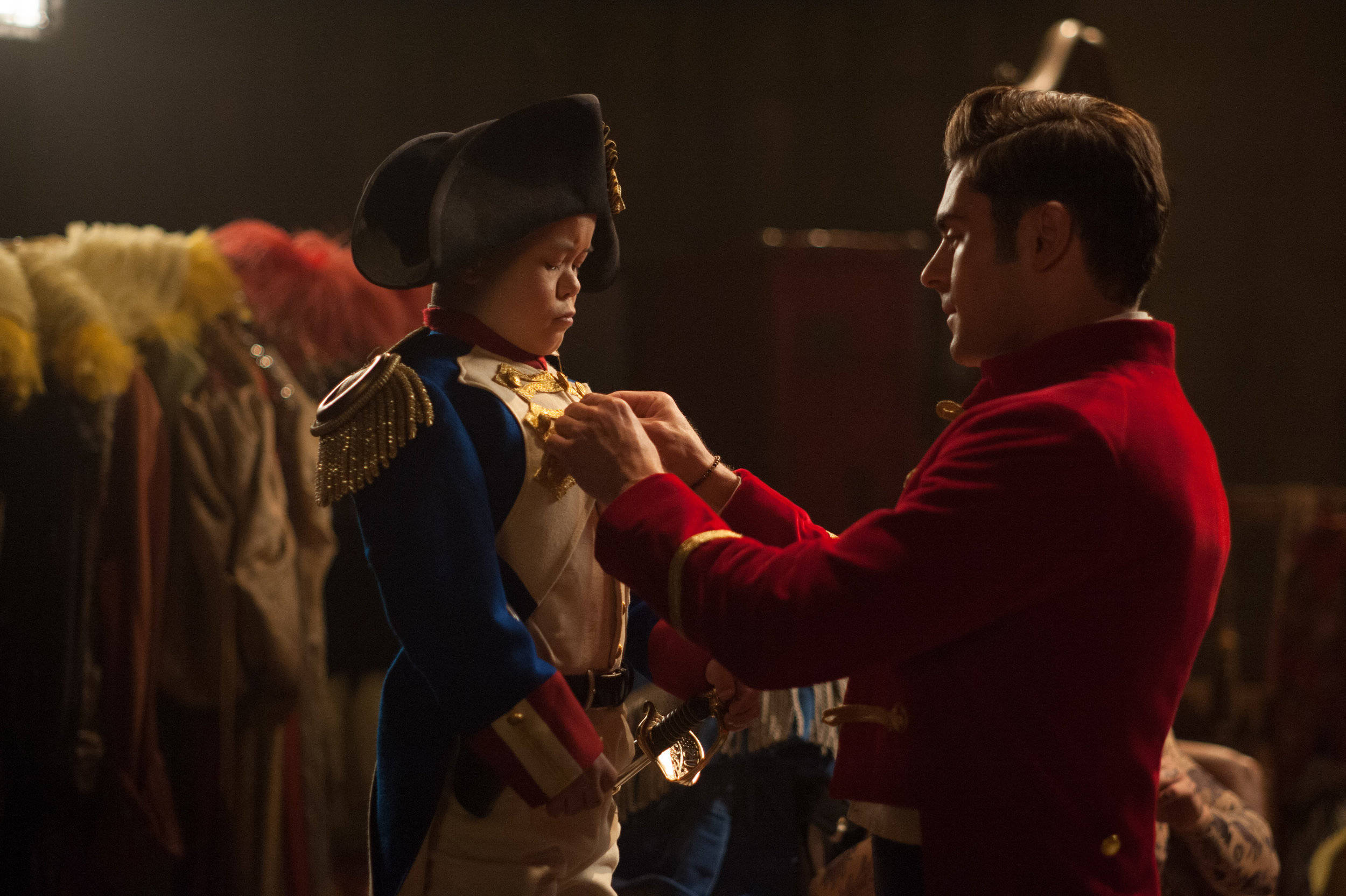 《 Greatest Showman 》黑暗場景 壁紙 — 一名身穿紅色外套的男子在溫暖的舞台燈光下，將一枚金牌別在一位身穿藍白軍裝的男孩身上。