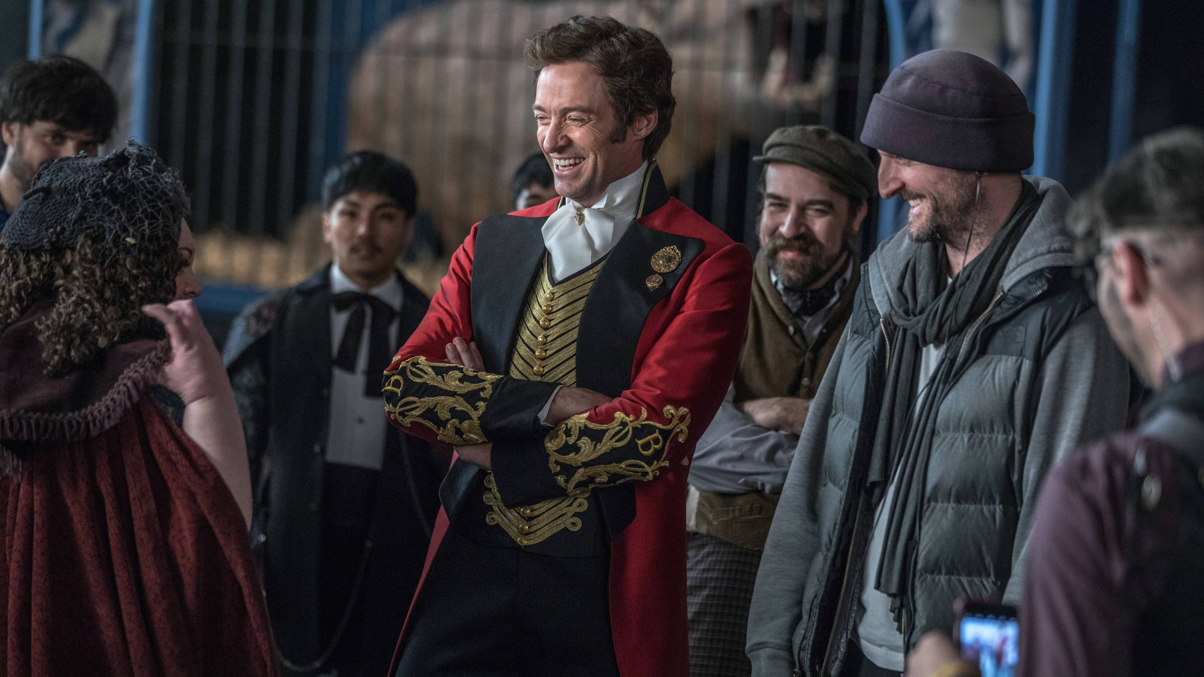 《 Greatest Showman 》歡樂時刻 壁紙 — 一名身穿紅金色戲劇服裝的男子在後台環境中被一群多元化的人包圍，開懷大笑。
