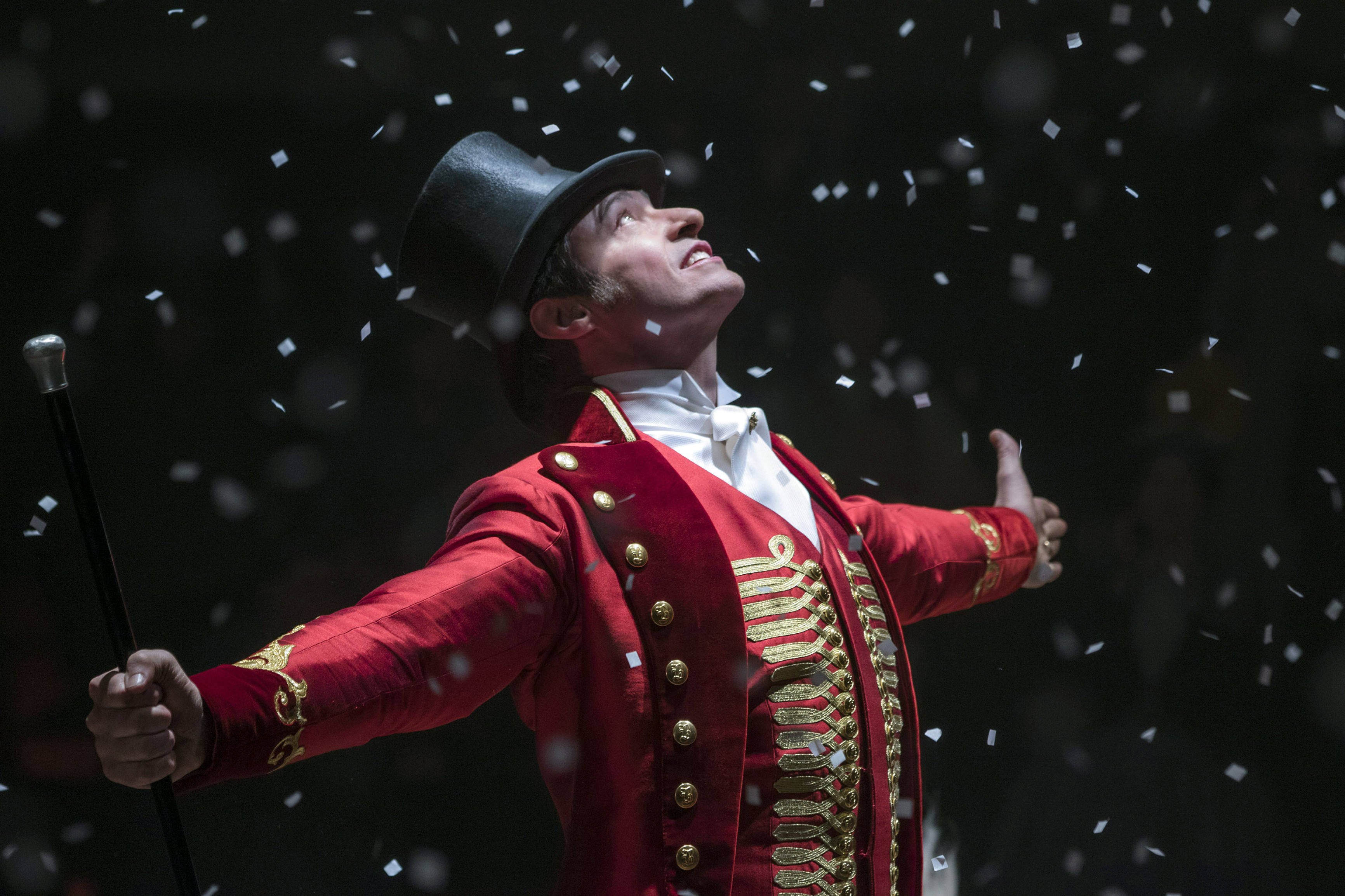 《 Greatest Showman 》主角休·傑克曼 壁紙 — 一位身穿紅金馬戲團服裝、戴著高帽的表演者在舞台燈光下歡快地向上拋灑五彩紙屑。
