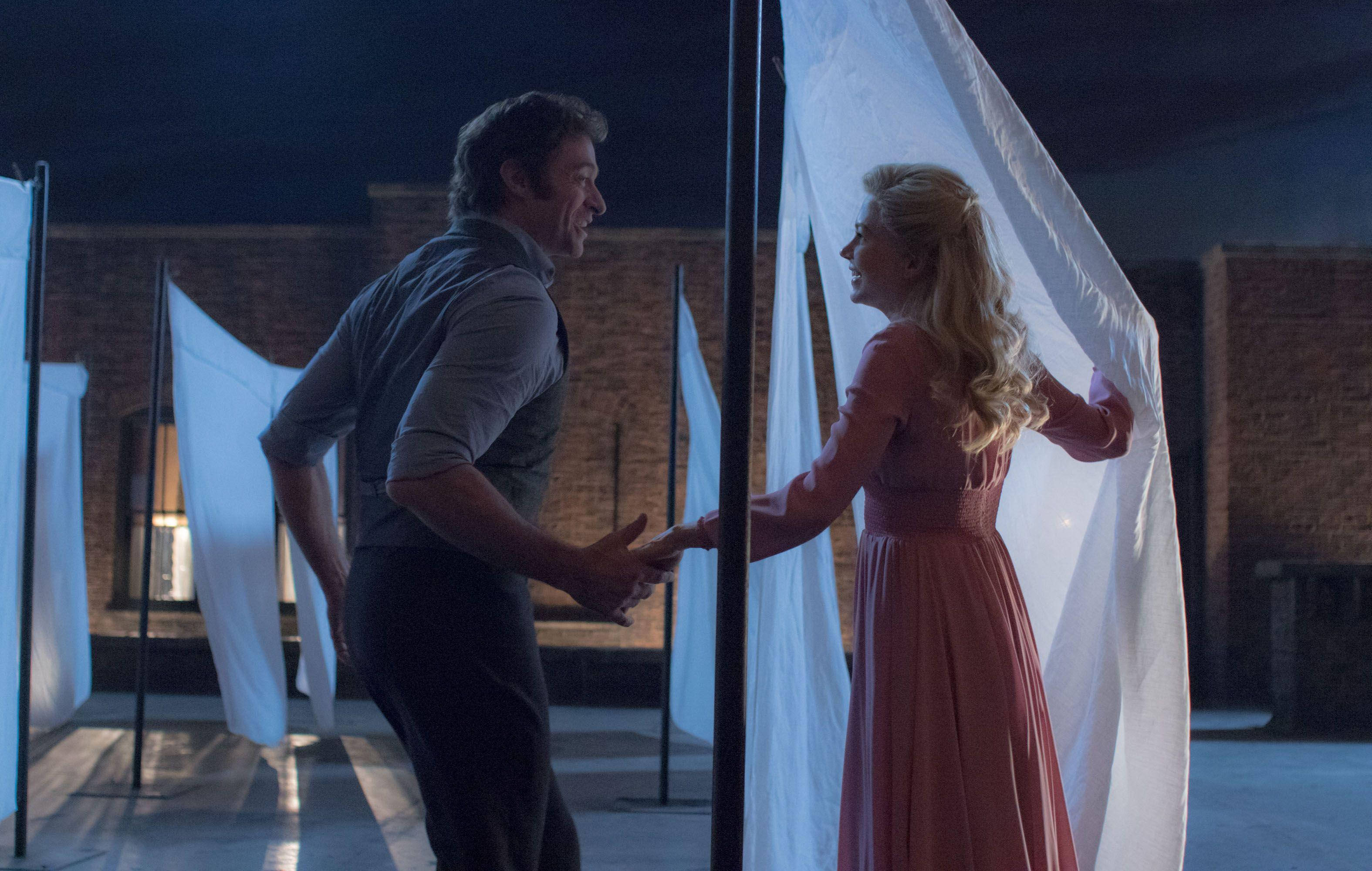 《 Greatest Showman 》巴納姆情侶 壁紙 — 夜晚，一男一女在昏暗的城市庭院中，在發光的白色布料面板下牽手。