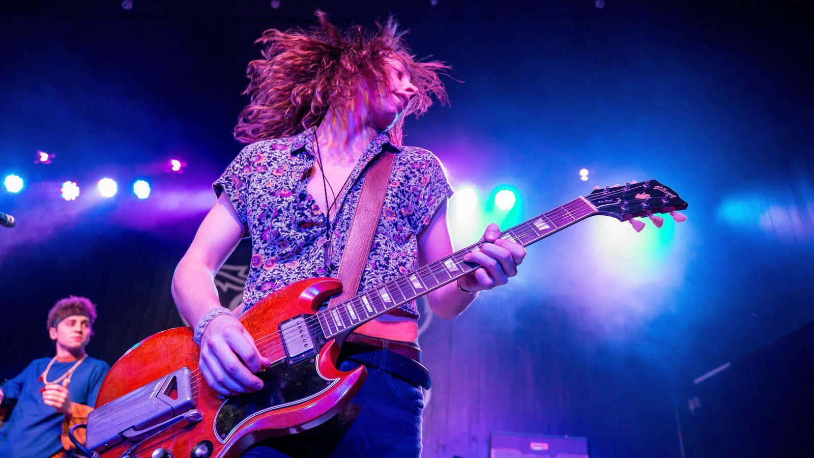 Greta Van Fleet 吉他手 Jake Kiszka 演唱會照 壁紙 — 一位擁有狂野紅髮的結他手，在充滿活力的藍色和紫色舞台燈光下，熱情地彈奏著一把紅色電結他。