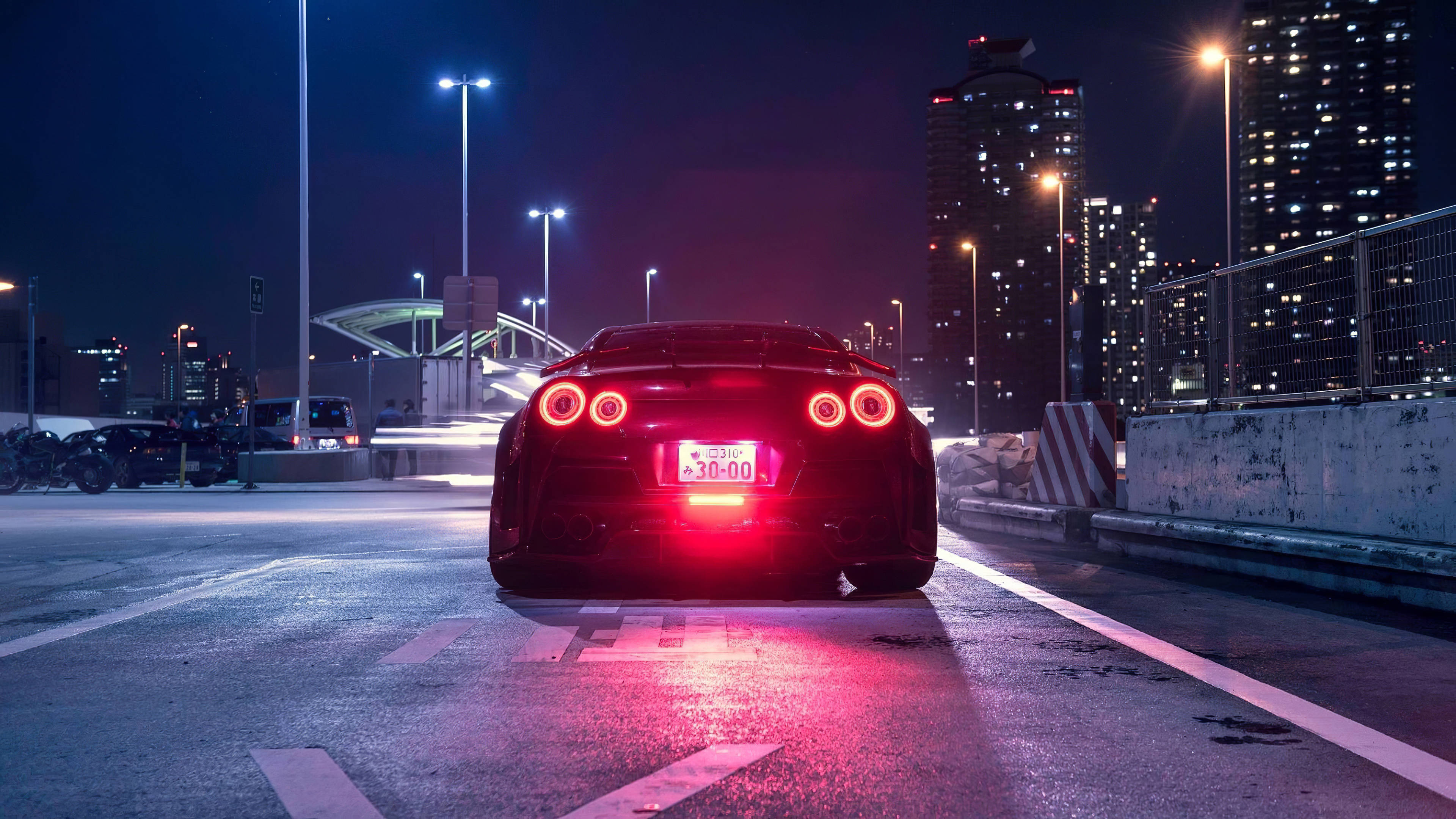 尾燈亮起黑色尼桑GT-R 4K 壁紙 — 一輛黑色跑車帶有發光的紅色尾燈，夜晚停在濕漉漉的城市道路上，周圍是高聳的燈光摩天大樓。