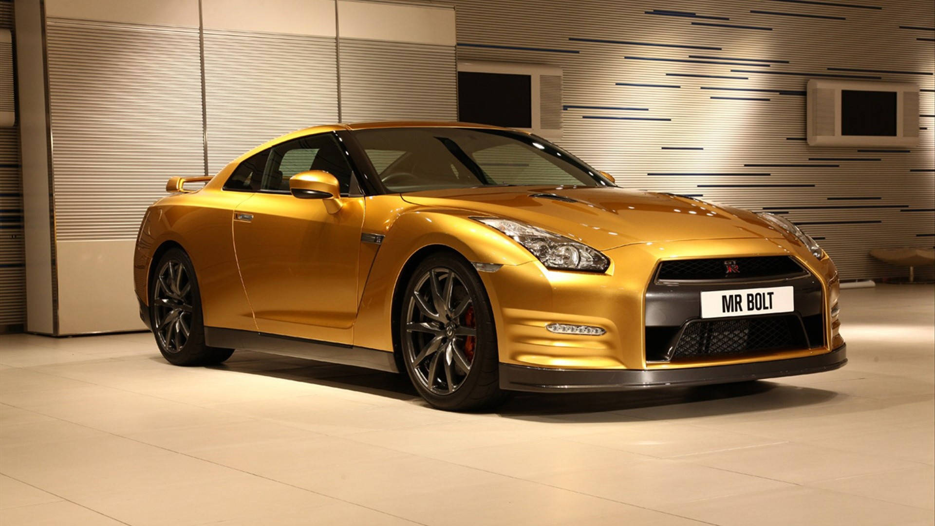 尼桑GT-R 華麗金漆 壁紙 — 一輛閃亮的金色日產GT-R跑車，帶有黑色飾件和紅色制動卡鉗，在一個現代簡約的室內展廳中展示，牆壁光滑，地板拋光。