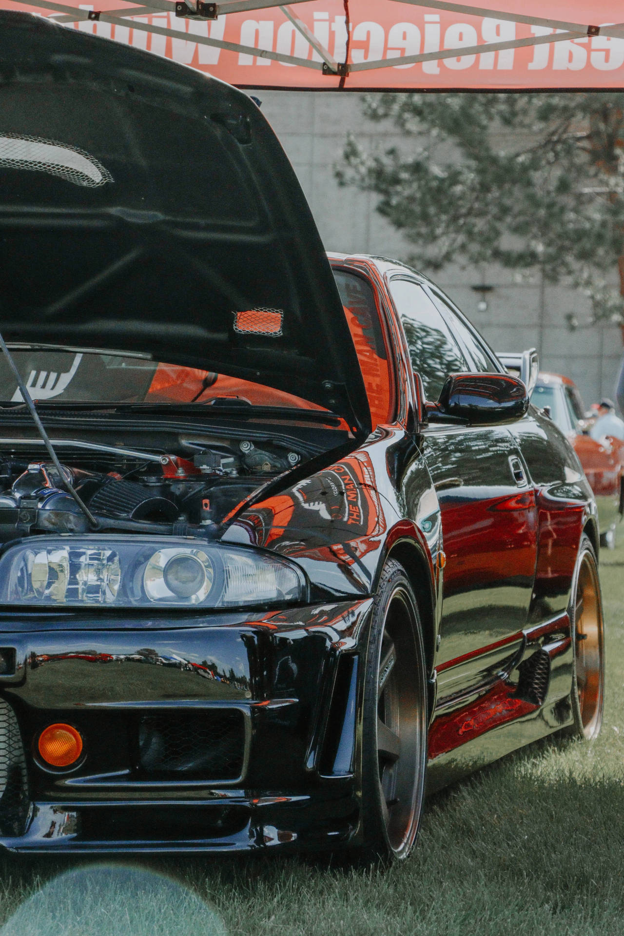 GT-R R33 車款引擎蓋打開 壁紙 — 一架亮黑色改裝跑車打開引擎蓋，在戶外車展的帳篷下展示紅色引擎。