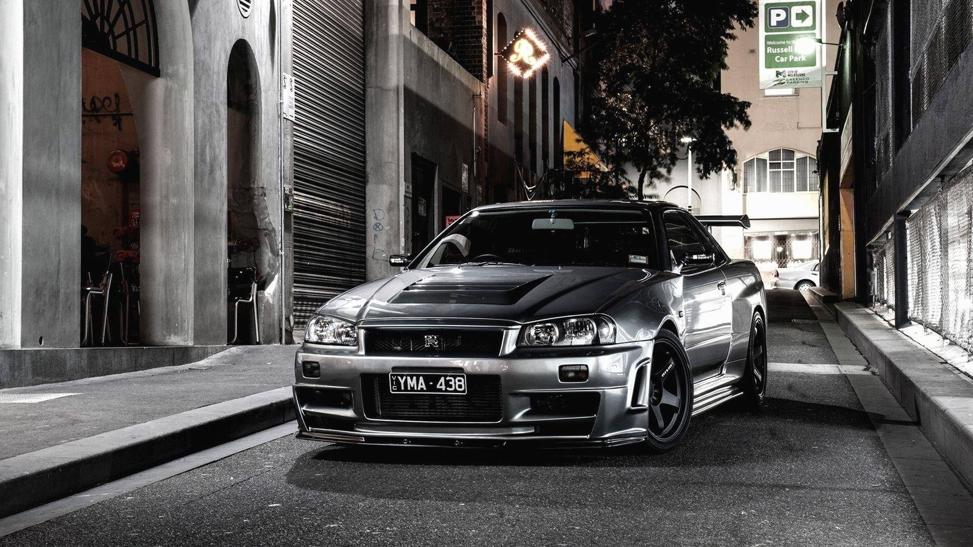 日產GT-R34全高清汽車壁紙 壁紙 — 一輛銀色日產Skyline R34跑車在夜間停泊在狹窄的城市小巷中，帶有戲劇性的燈光。