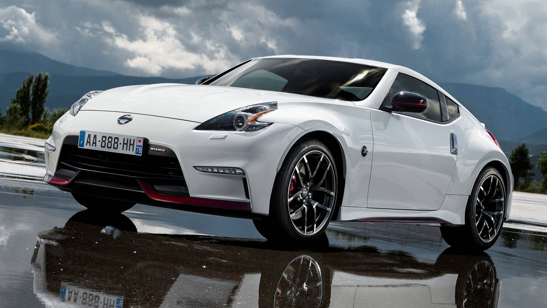 日產GT-R白Nismo 壁紙 — 一架時尚的白色日產370Z跑車，配備黑色輪圈和紅色飾邊，停在潮濕的道路上，在陰天和山脈背景下反射其影像。