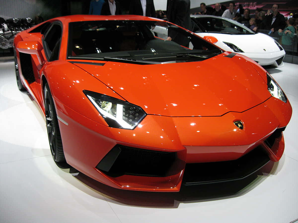 一輛蘭博基尼GT4在室內展示 壁紙 — 一輛鮮橙色的林寶堅尼Aventador在車展的明亮燈光下閃閃發光，背景有一輛白色的林寶堅尼。