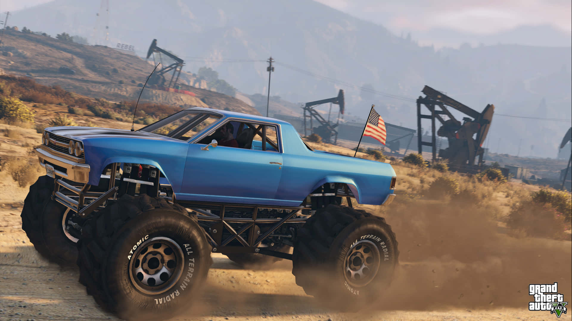 GTA 5 藍色怪獸卡車背景 壁紙 — 一輛藍色怪獸卡車用超大輪胎揚起灰塵，在點綴著油泵的乾燥沙漠景觀中疾馳。
