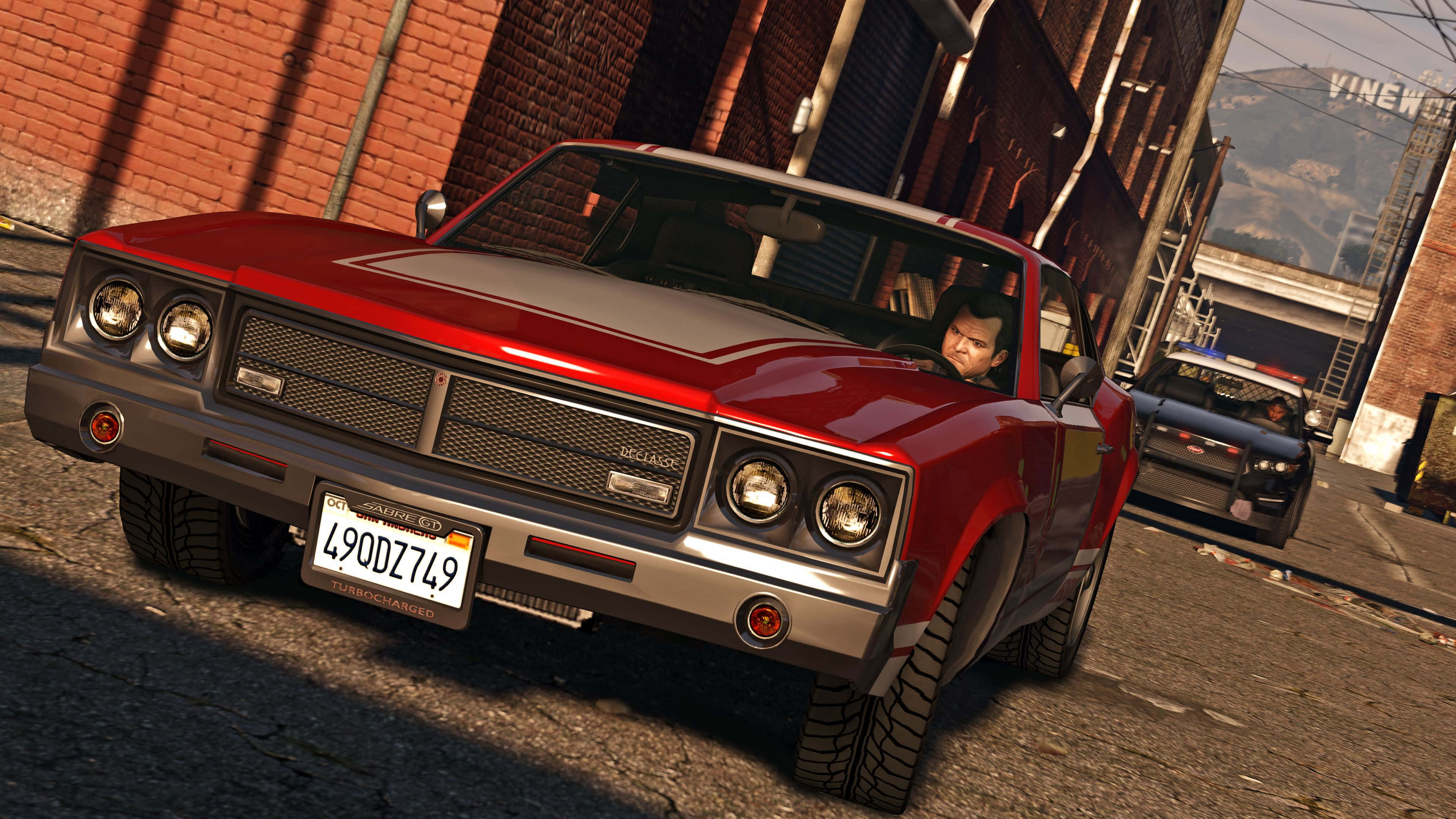 GTA V 4K風景 壁紙 — 一輛帶有鍍鉻細節的紅色經典肌肉車在磚砌建築之間的陽光普照的城市小巷中行駛。