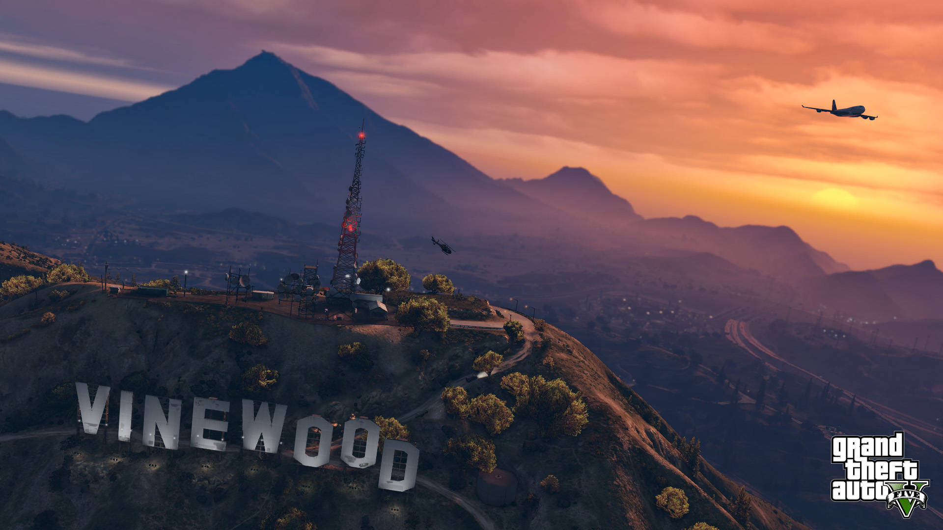 GTA V 文森伍德山 壁紙 — 山巒景觀上的日落，有一個大型「VINEWOOD」標誌、一座無線電塔和一架飛機在頭頂飛過。