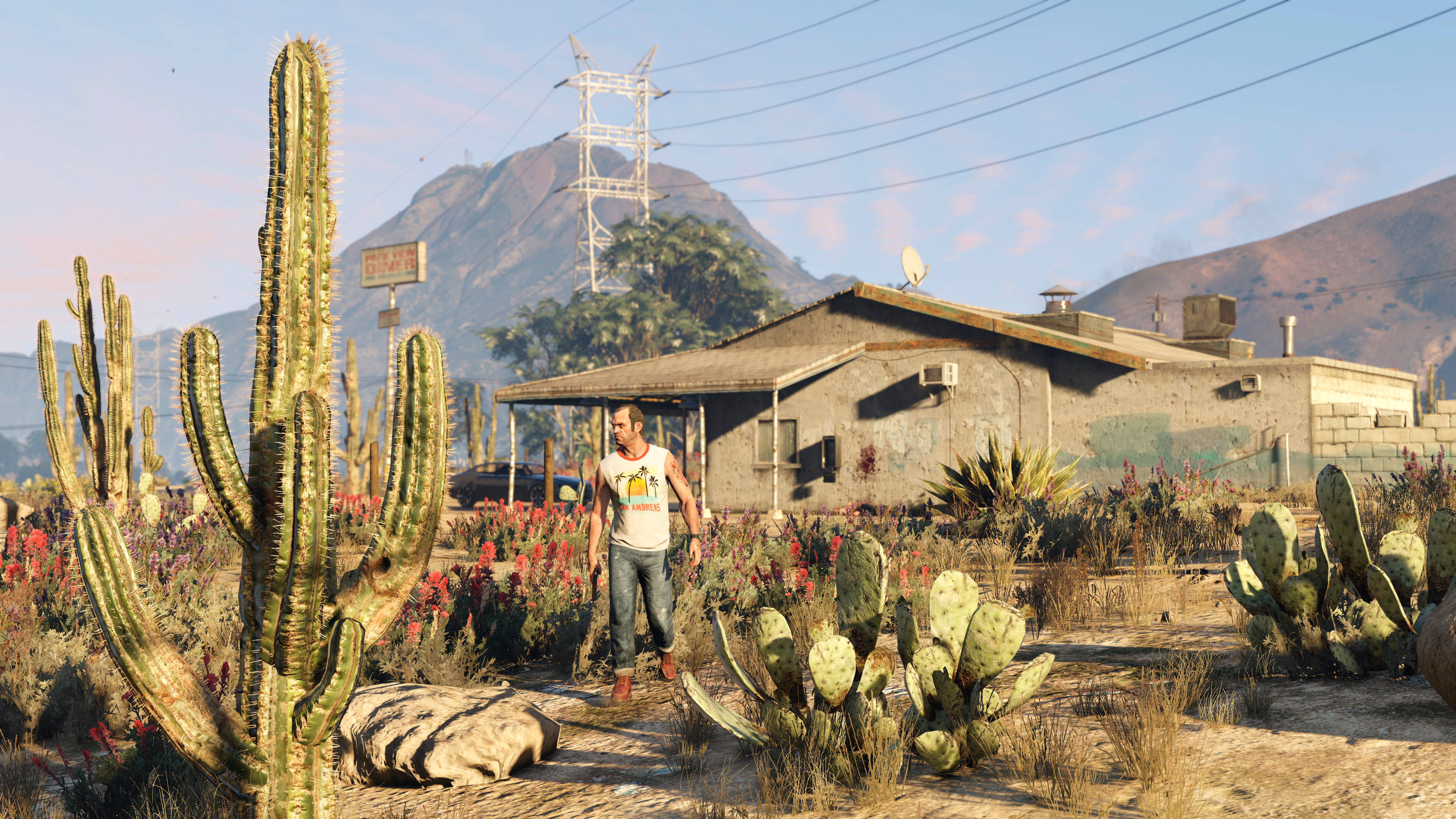 GTA V 特雷弗沙漠植物 壁紙 — 一個男人在晴朗的藍天下走過沙漠景觀，有仙人掌和一座風化的房子。