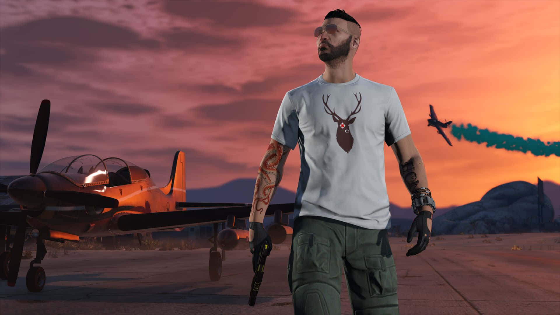 GTA紋身男子角色圖 壁紙 — 一個穿著鹿紋T恤的男人站在一架復古飛機旁邊，而一架噴射機在沙漠日落時疾馳而過。