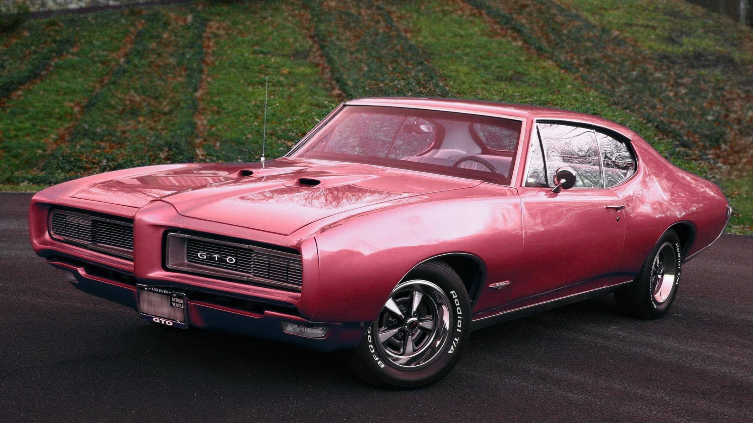 高畫質經典粉紅龐蒂克GTO 壁紙 — 一架閃亮粉紅色1960年代Pontiac GTO肌肉車停在瀝青路上，背景有草地山坡。