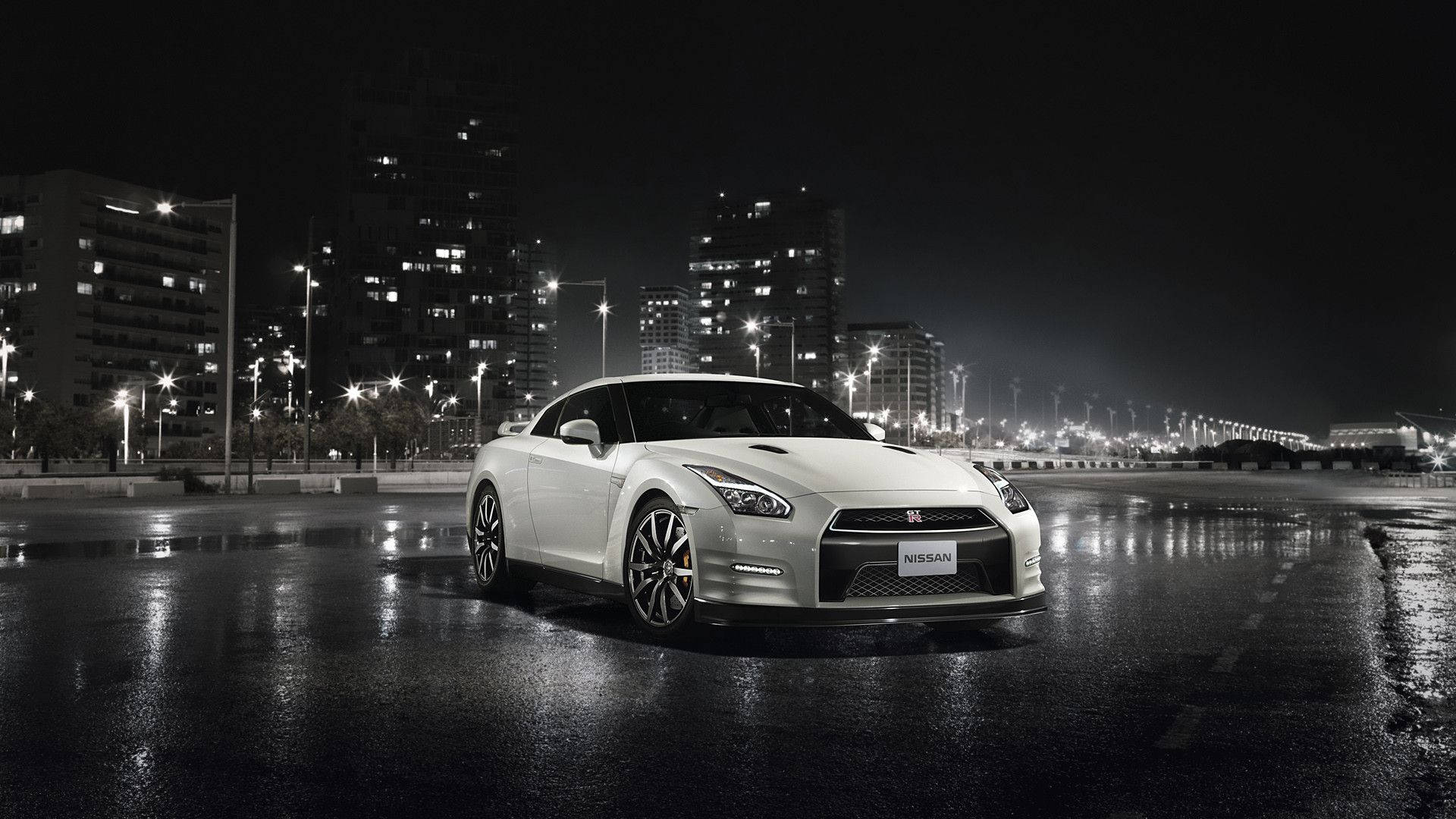 城市夜景中的白色日產GTR跑車 壁紙 — 一輛流線型白色日產GT-R在夜間濕漉漉的瀝青路上，於城市燈光下閃耀，背景是高樓大廈發出的光芒。
