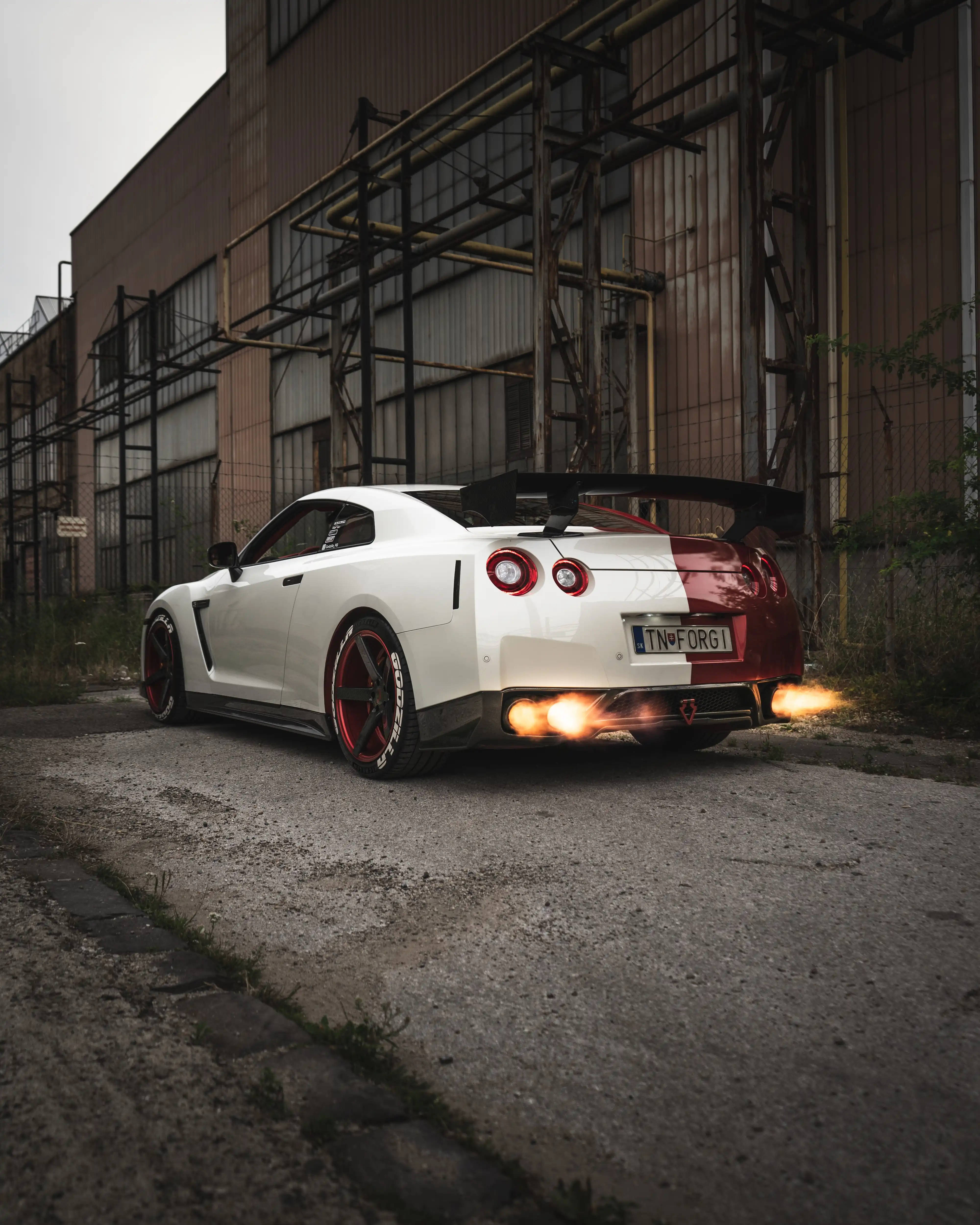 GTR 車款上的迷人藝術創作 壁紙 — 一輛白色和紅色的日產GT-R，帶有發光的排氣管，停在生鏽的工業建築前。