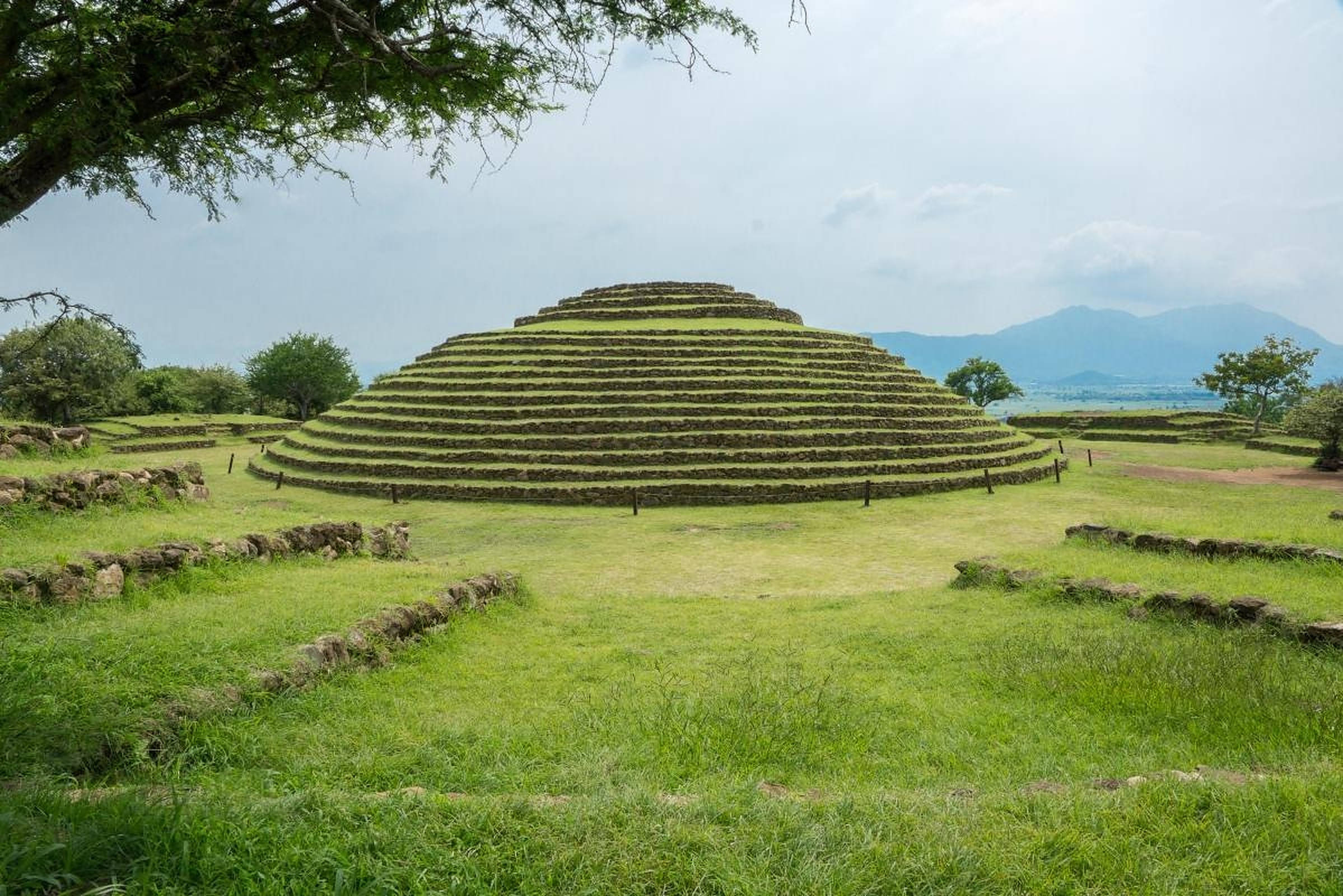 瓜達拉哈拉Guachimontones金字塔 壁紙 — 一座巨大的古老階梯金字塔，覆蓋著充滿活力的綠草，矗立在寧靜的開闊田野中，天空多雲，遠處有山脈。
