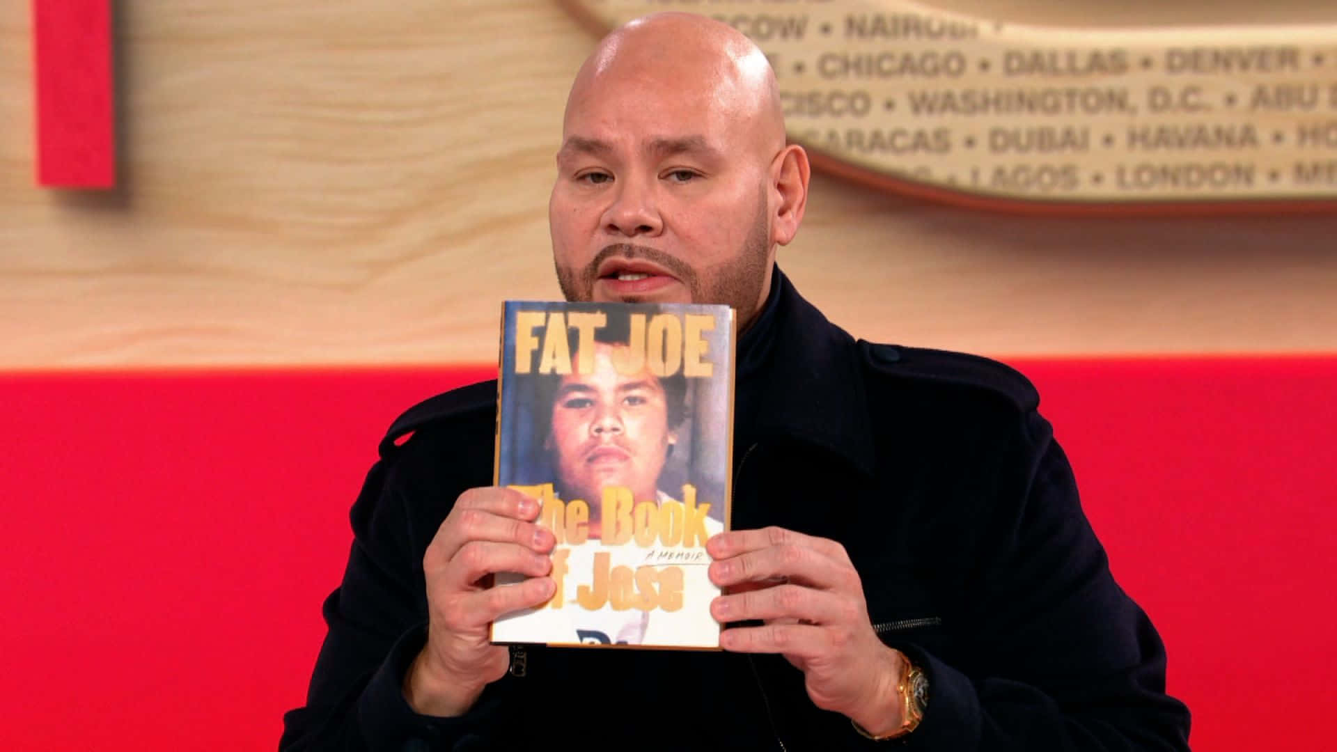  壁紙 — 光頭男子身穿黑色外套，在紅色和木紋背景前舉起一本名為《Fat Joe The Book of Jose》的書。