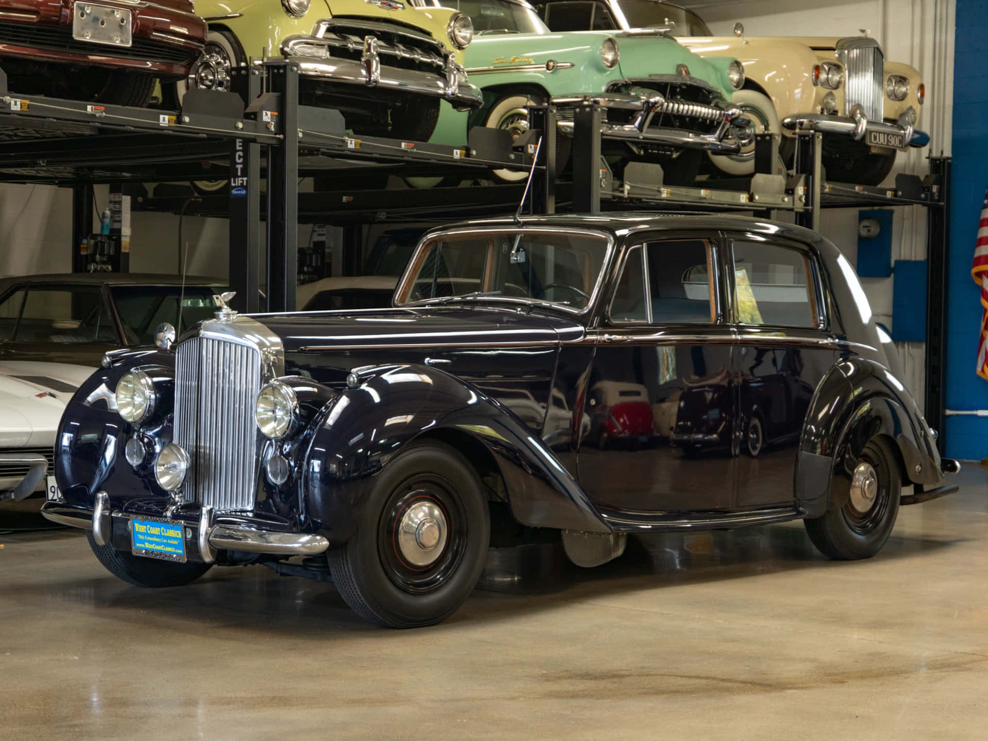 Bentley Mark Vi 桌布 壁紙 — 一架光澤的海軍藍色 vintage Rolls Royce 在光線充足的車庫中突出地停放，後面有其他經典汽車堆疊在升降機上。